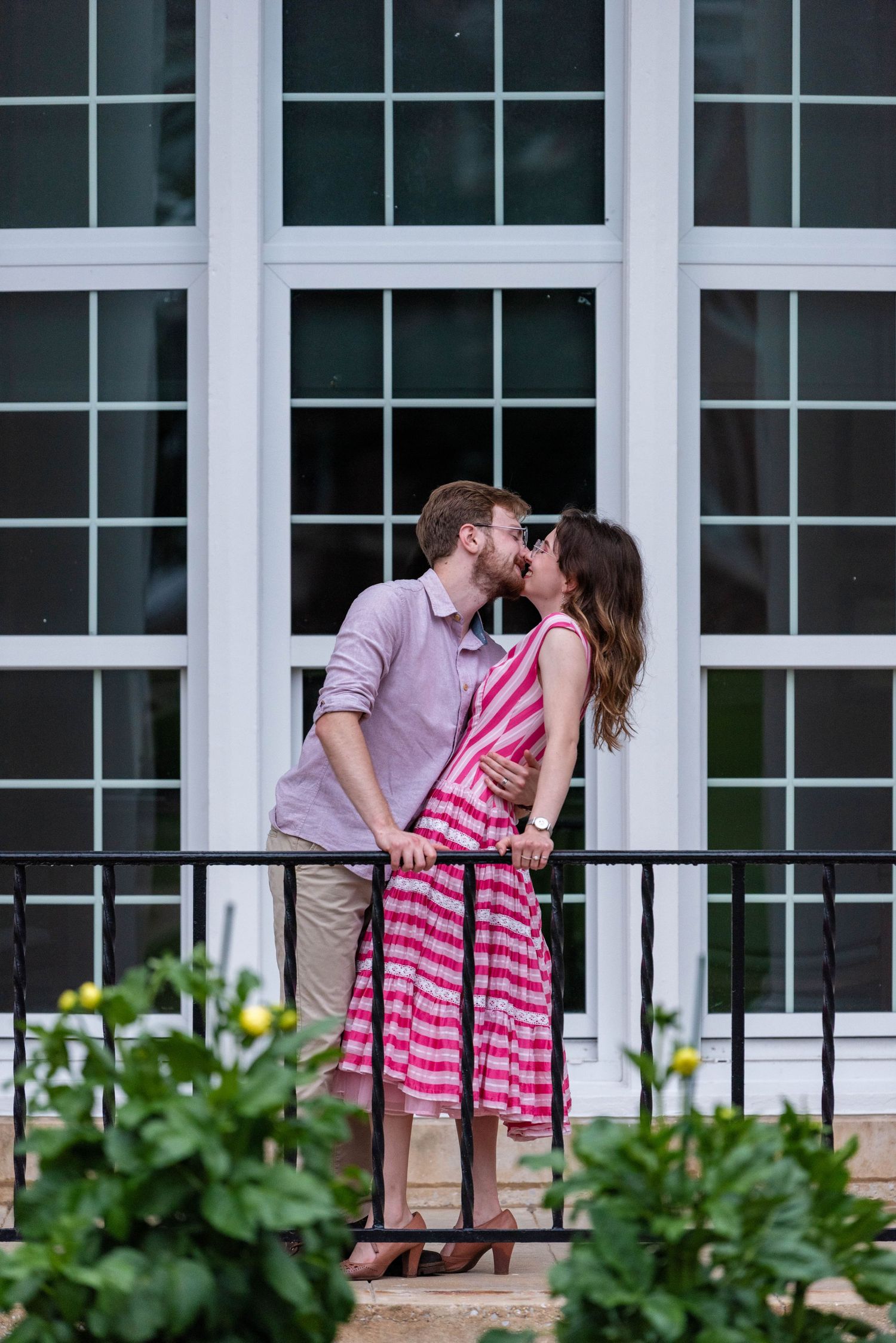 Engaged couple kissing on a porch with white French windows, woman wearing a pink striped dress.