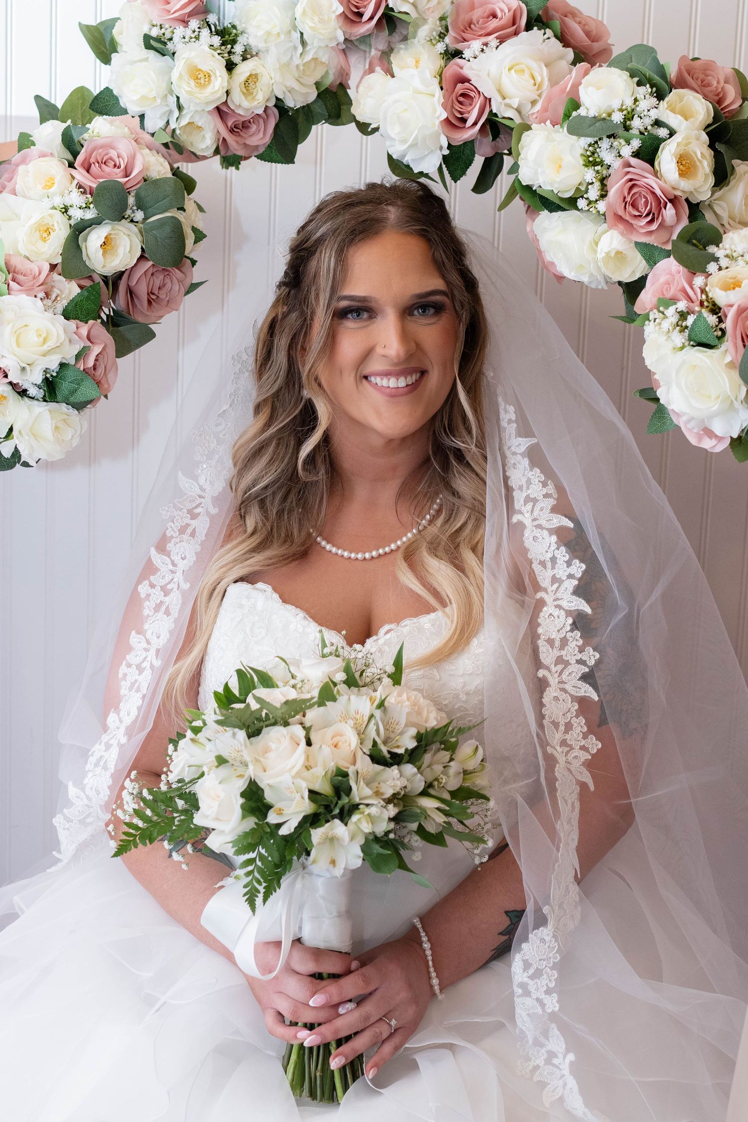 Smiling bride in lace veil holds white bouquet under pink and white floral arch.