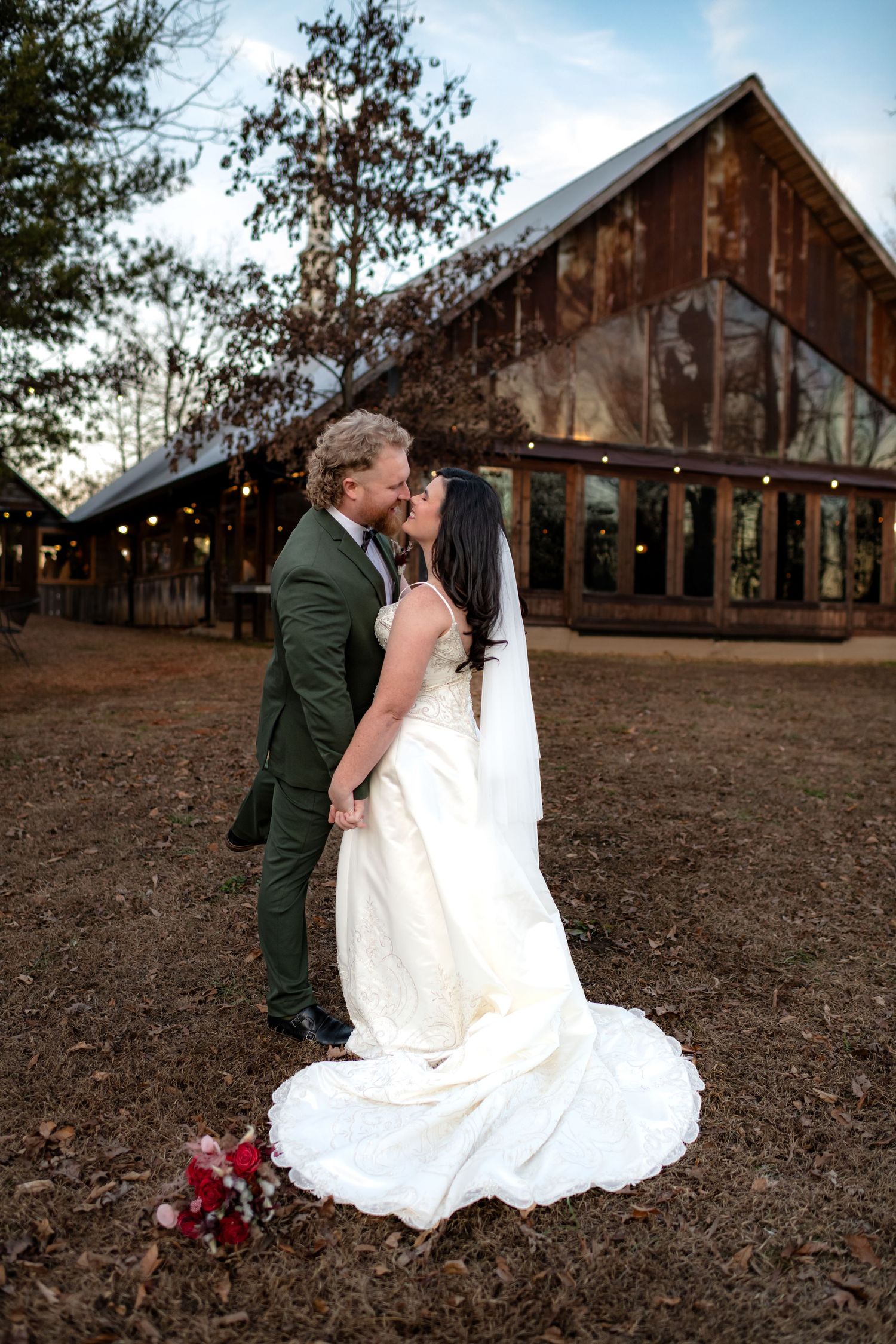 Bride and groom share a romantic kiss outside a rustic barn venue decorated with string lights at dusk.