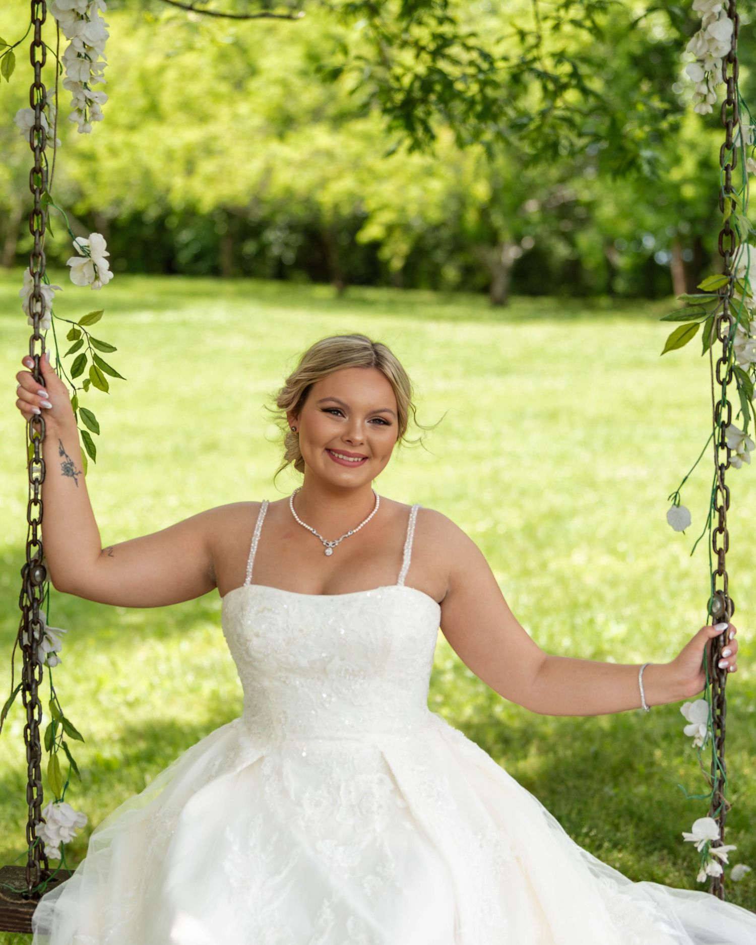 Smiling bride in white lace gown sitting on a floral chain swing surrounded by lush green trees.