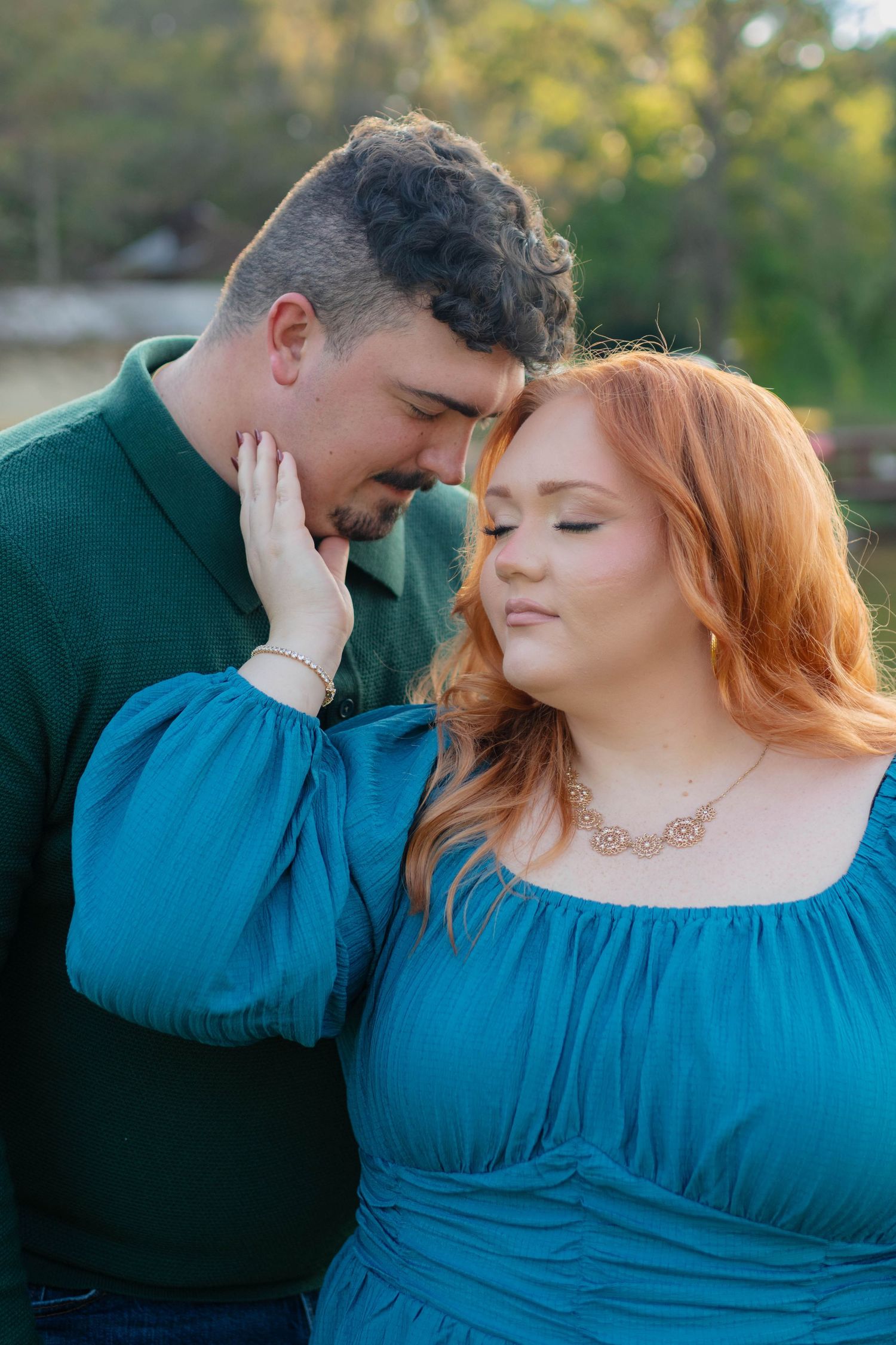 Romantic couple in teal and green touching foreheads tenderly during golden hour outdoors.