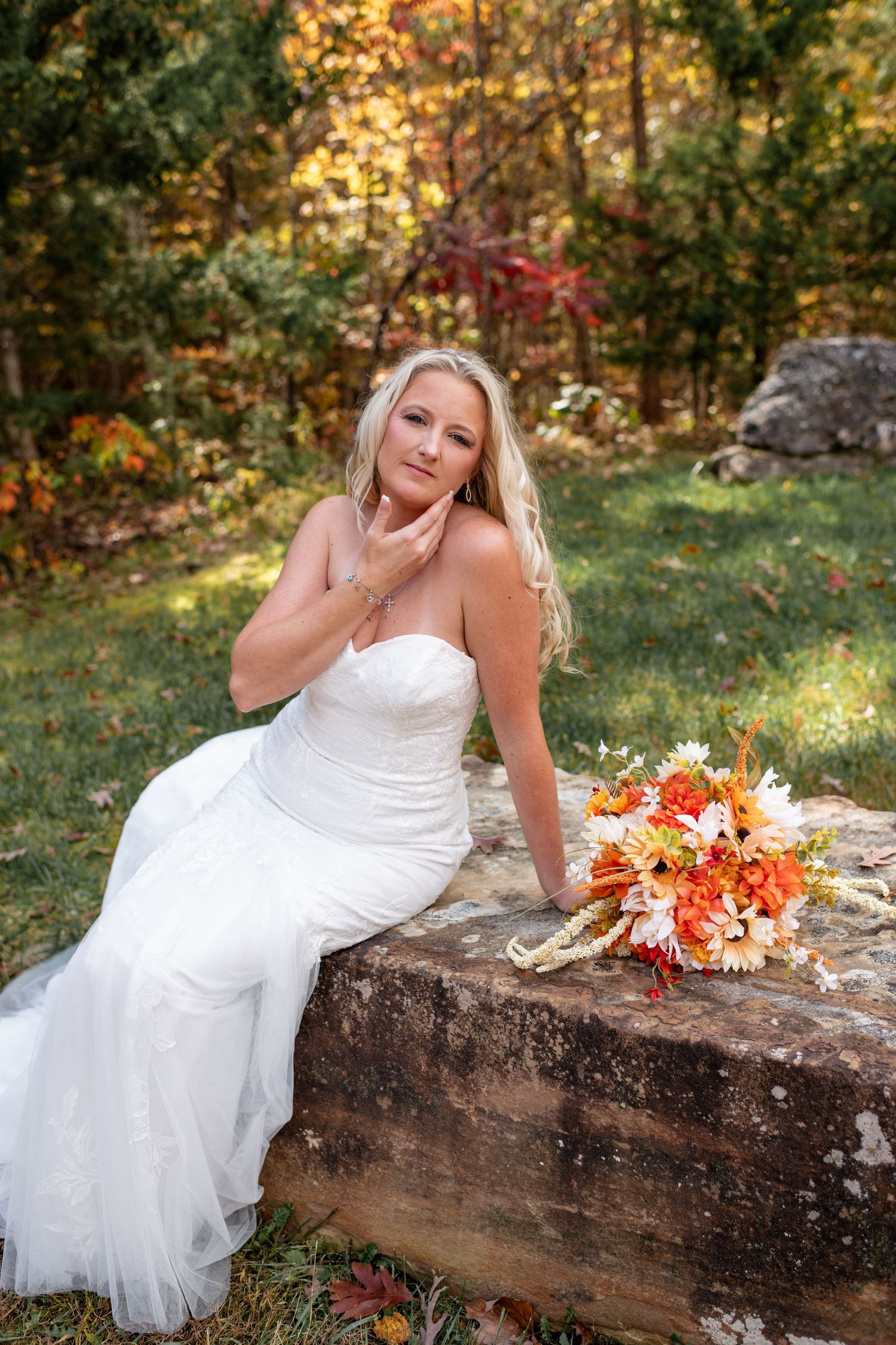 Elegant bride in strapless white gown sits on stone wall with orange fall bouquet in autumn forest.