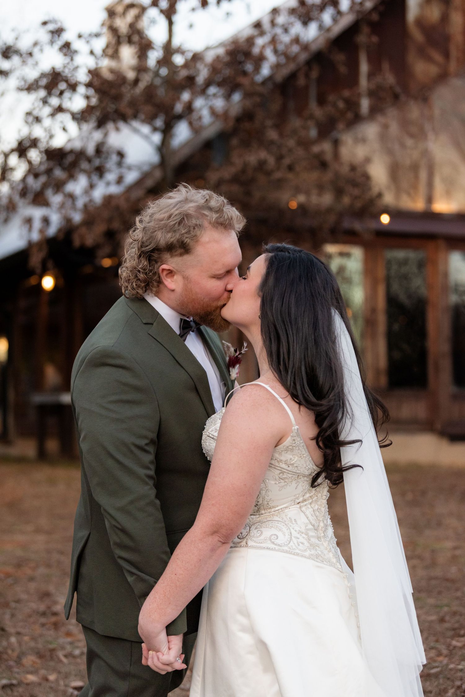 Bride and groom share a romantic kiss outside a rustic barn venue at dusk.