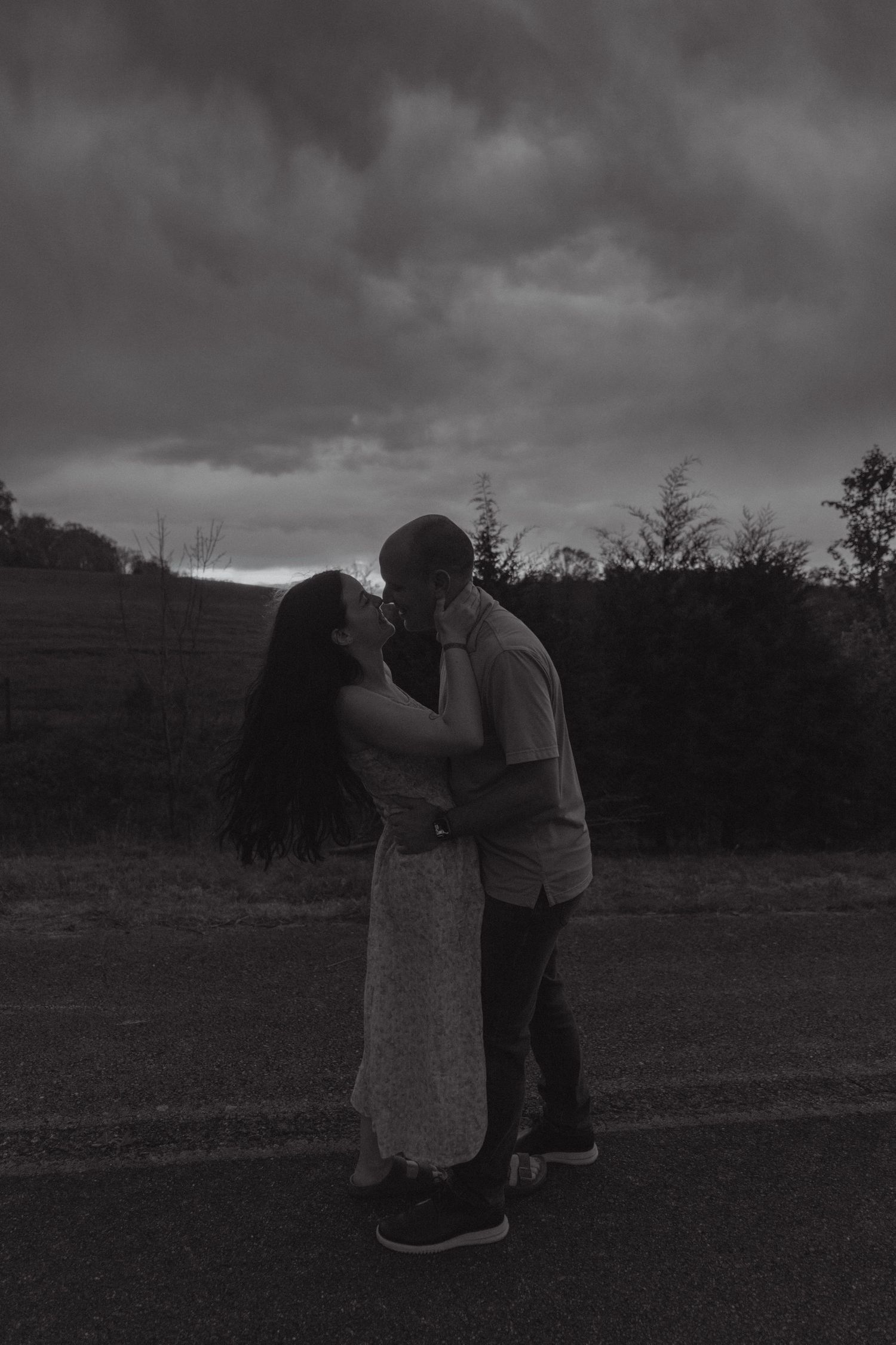 Black and white photo of a couple kissing on a road with dramatic cloudy sky and trees in background.