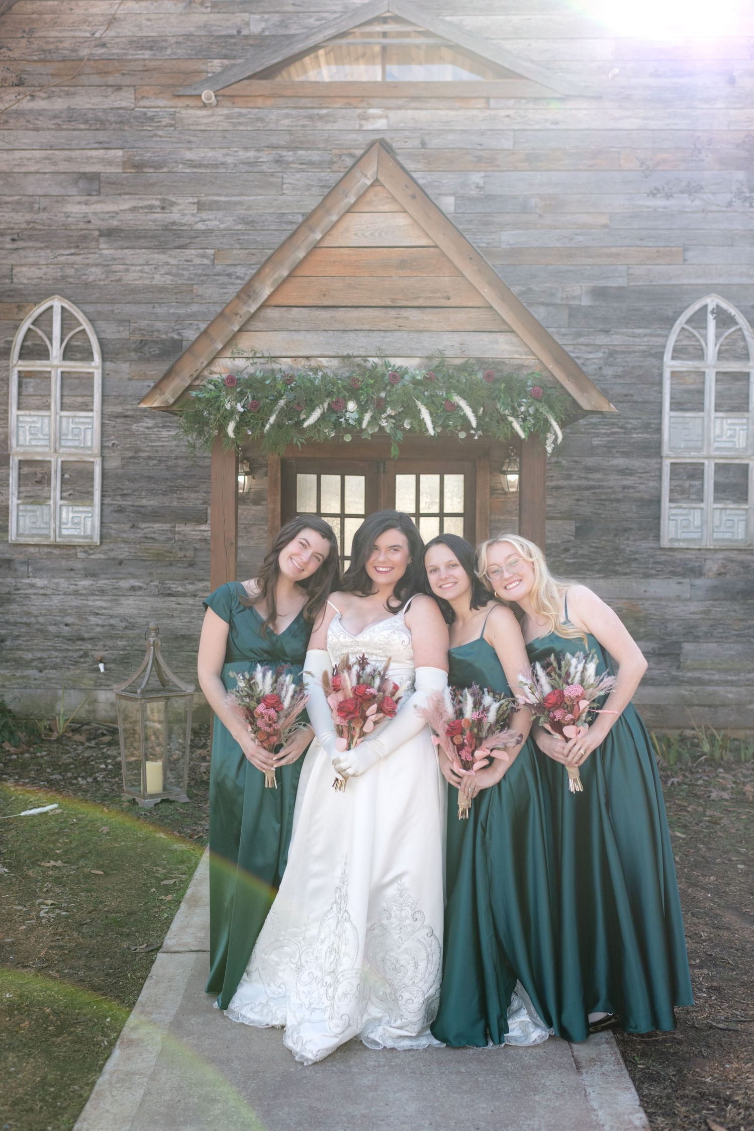 Bride with three bridesmaids in emerald gowns pose before a rustic wooden chapel entrance.