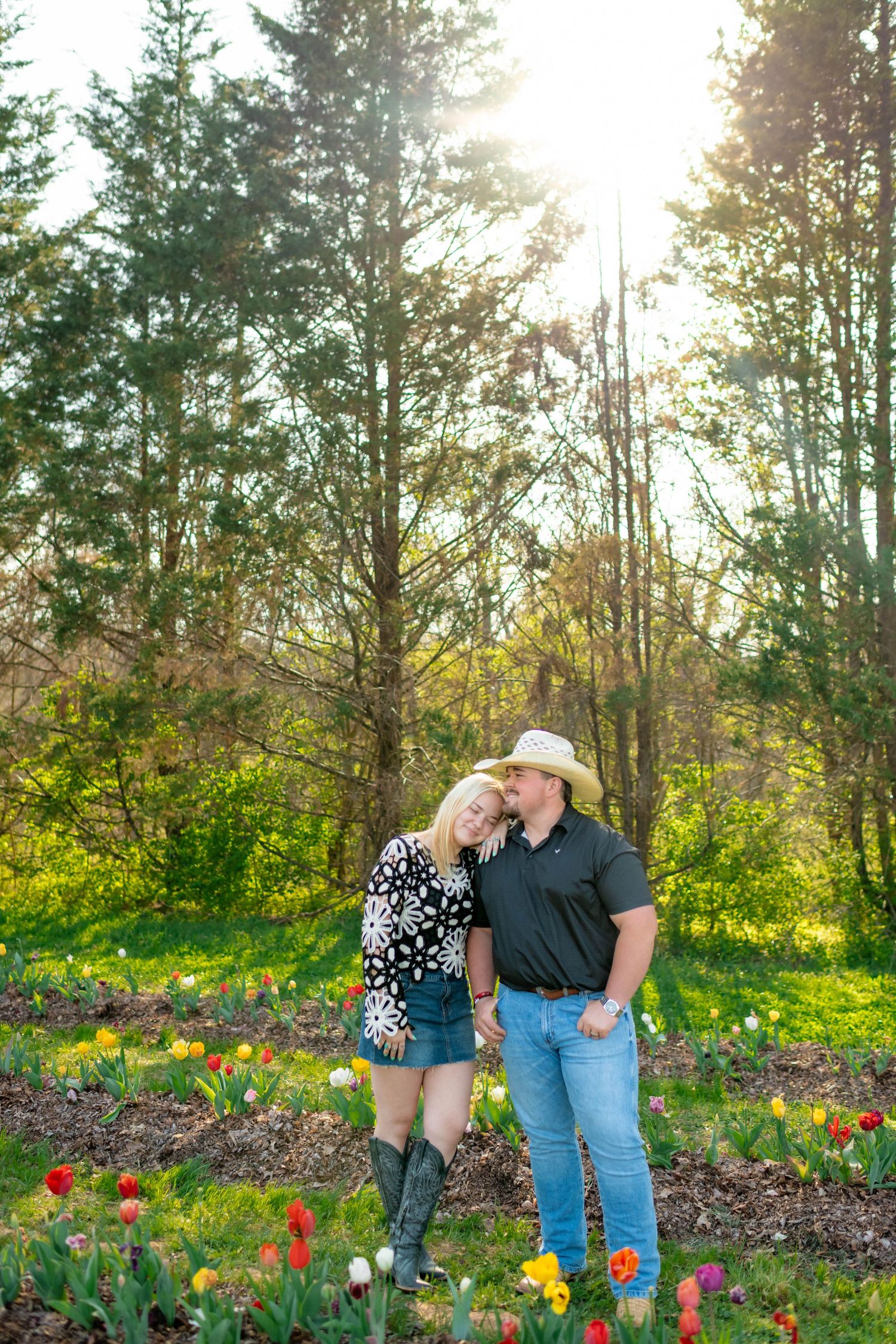 Couple embracing in a tulip garden with sunlight streaming through tall pine trees.