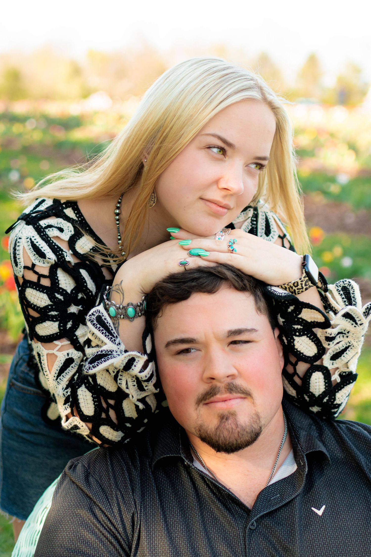 Blonde woman resting chin on mans head while posing together in a colorful outdoor garden setting.