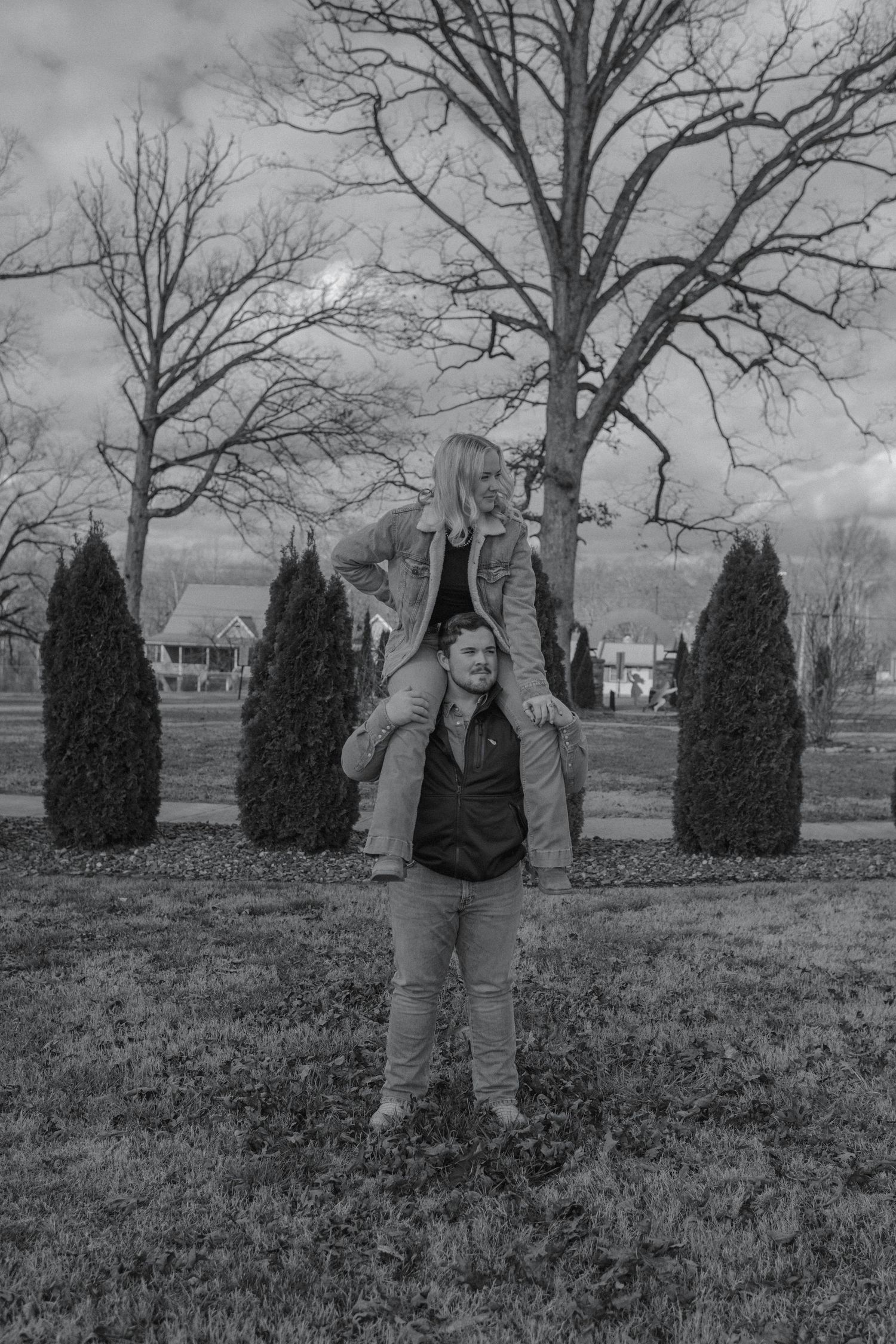 Black and white photo of a woman sitting on a man's shoulders in a park with bare trees and evergreen shrubs.