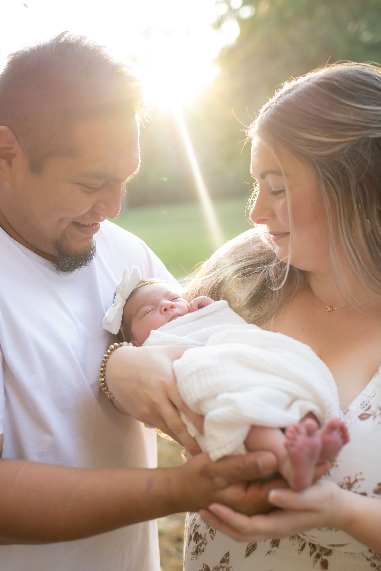 New parents tenderly cradling their swaddled newborn baby together in a warmly backlit outdoor setting.
