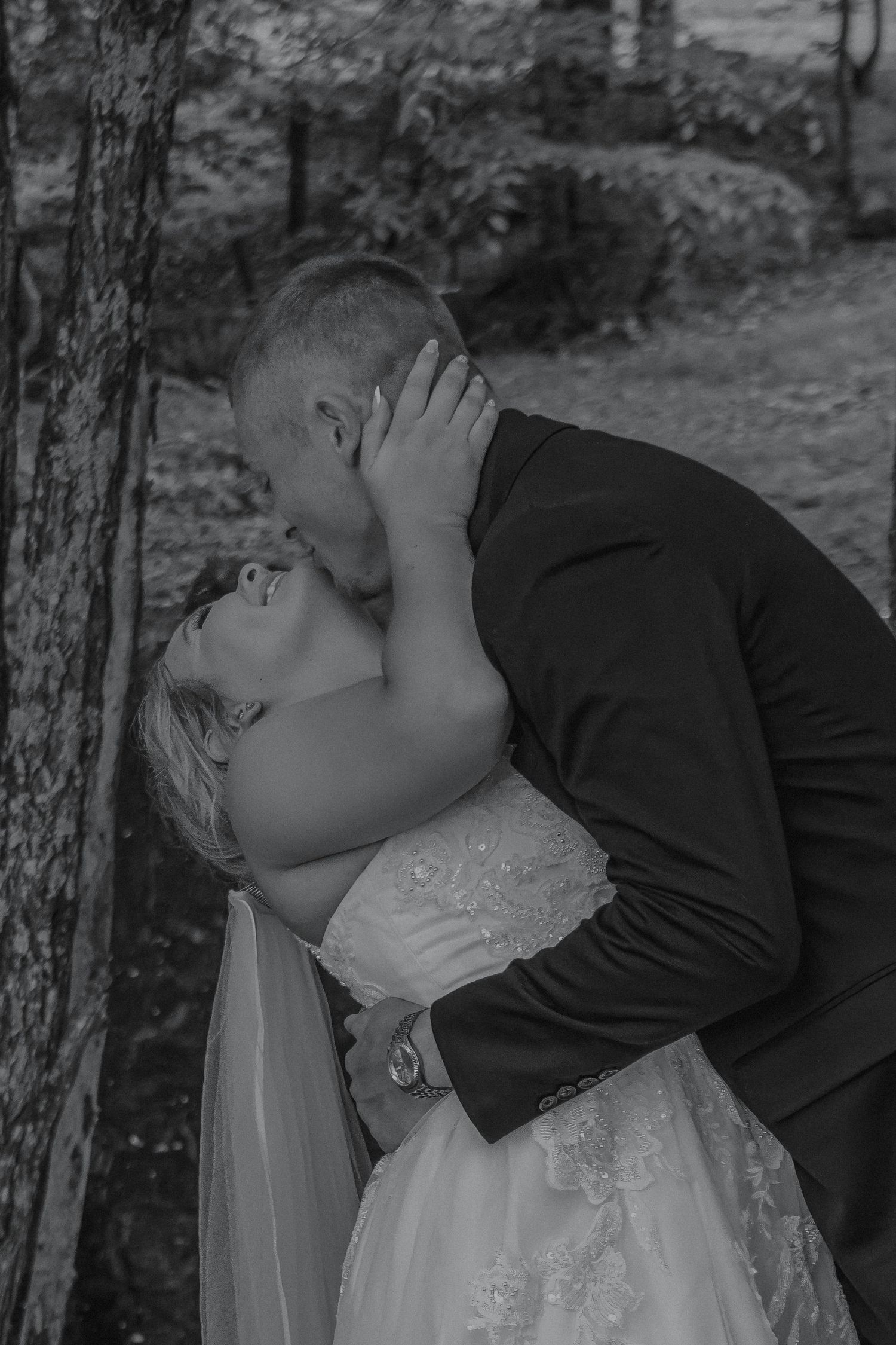 Black and white romantic photo of bride and groom sharing a passionate kiss beside a tree.