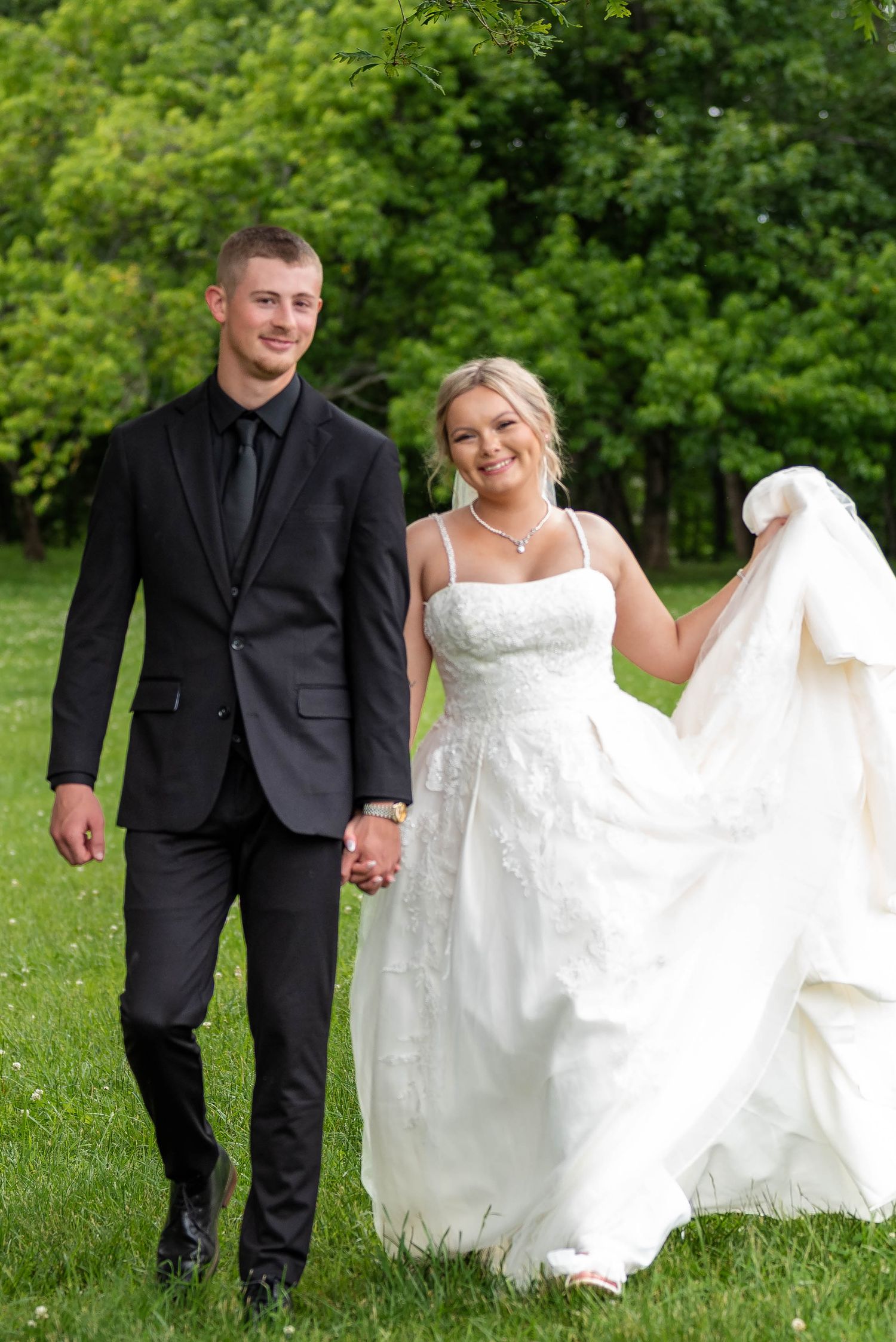 Smiling bride and groom walking hand in hand on green grass, bride lifting white ballgown.