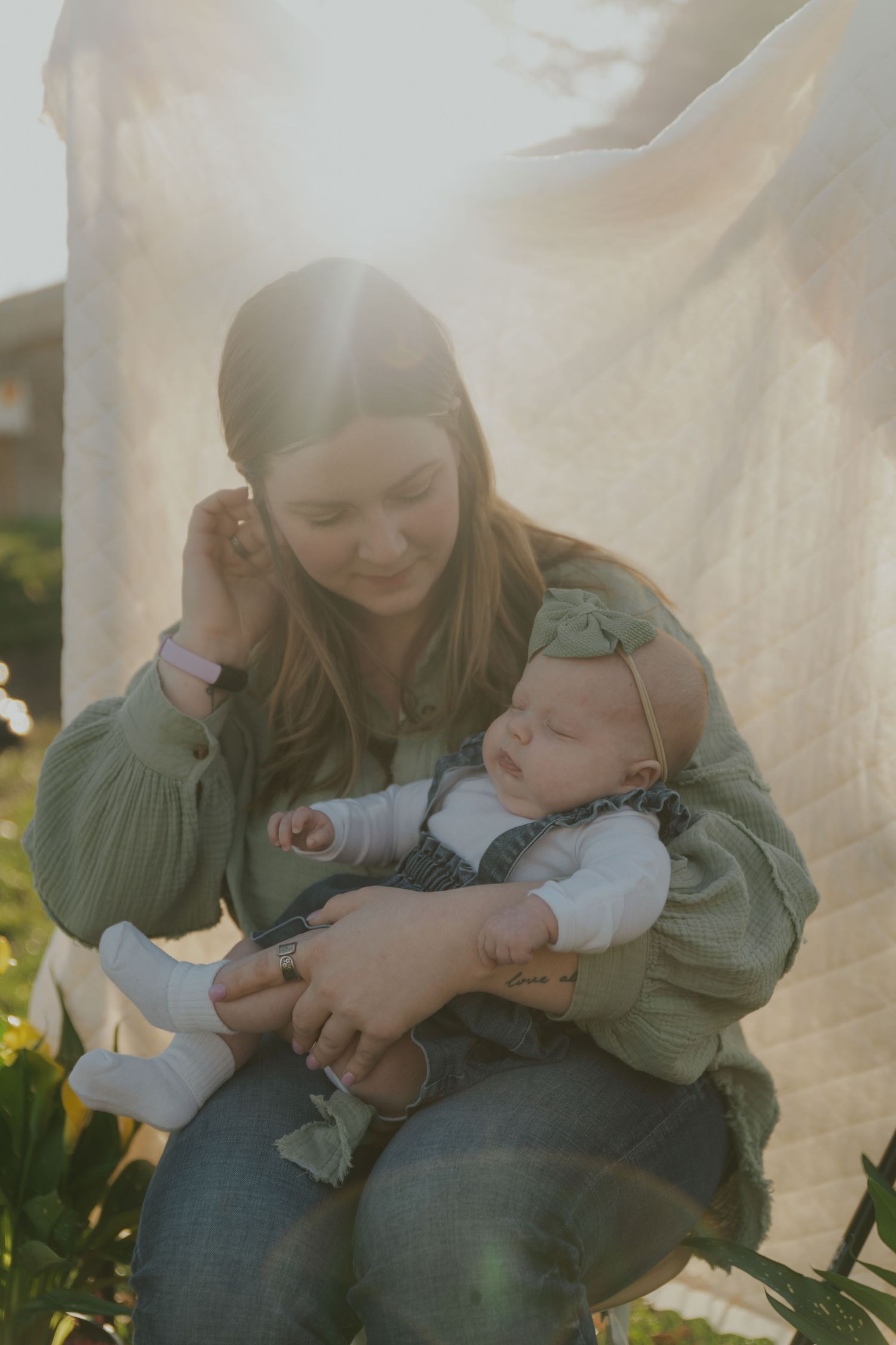 Young mother holding sleeping newborn baby in green bow headband during outdoor golden hour session.