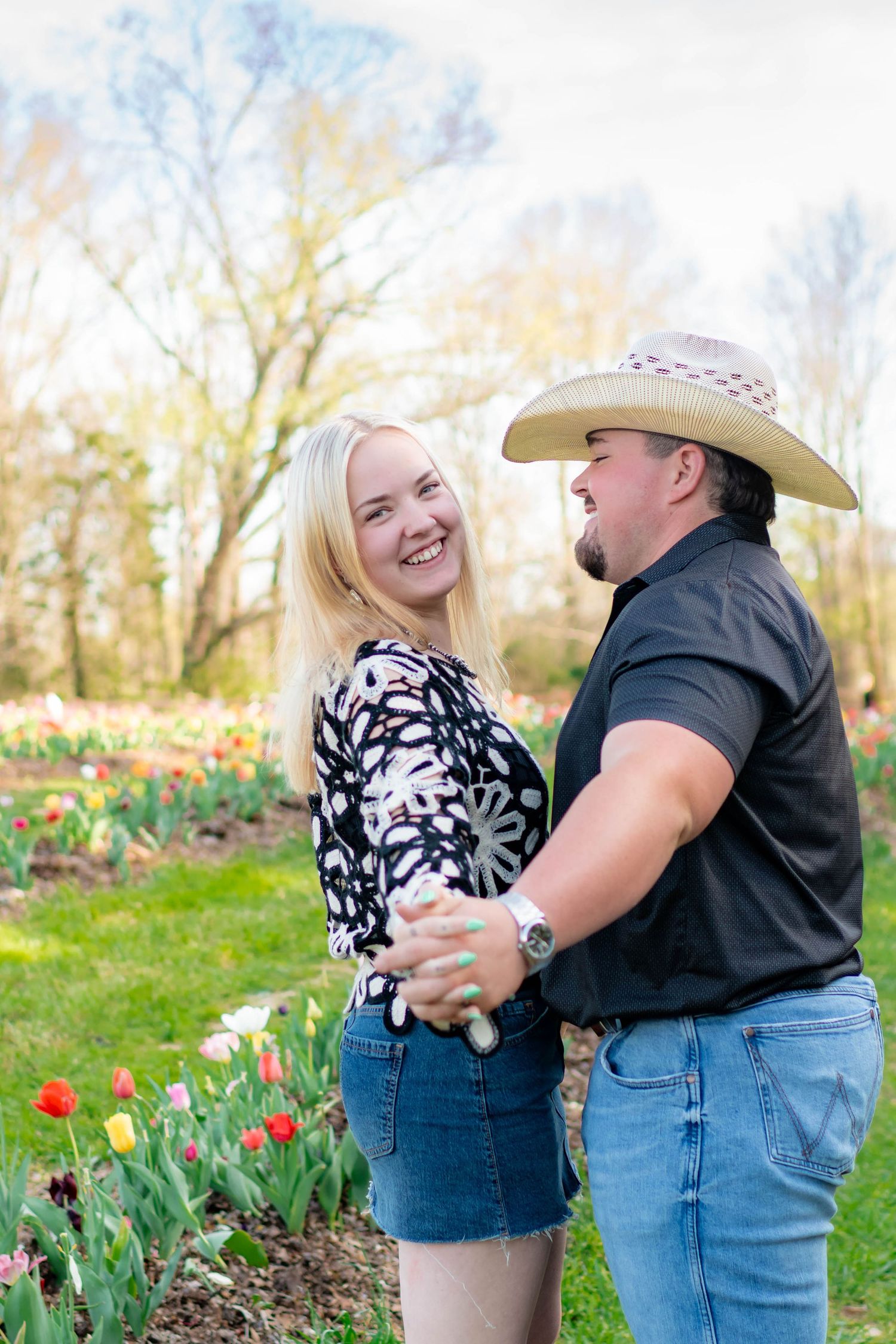 Smiling couple holding hands in tulip garden, man wearing cowboy hat, woman in floral top.