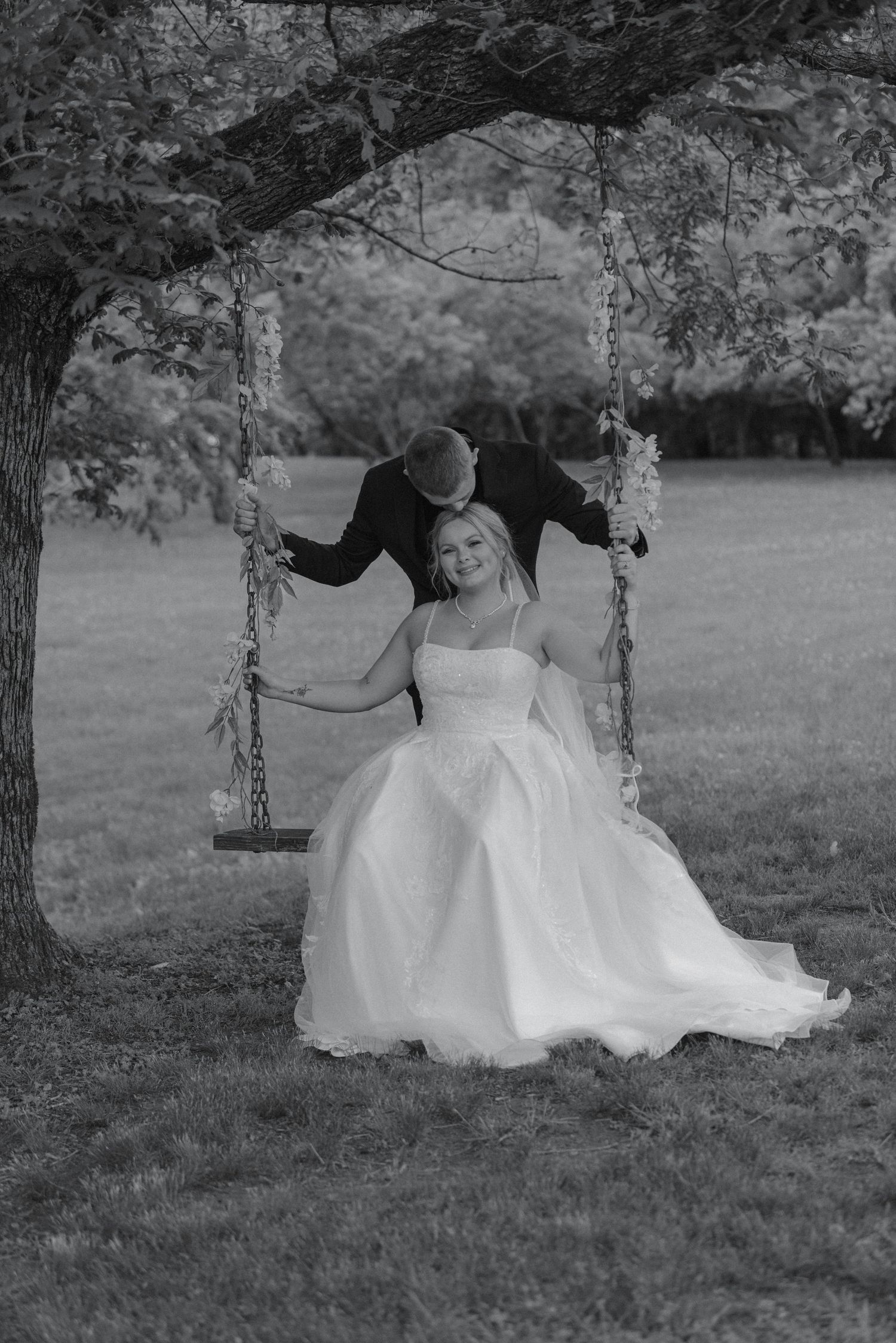 Black and white photo of groom kissing bride on a garden swing, bride in elegant white ballgown.