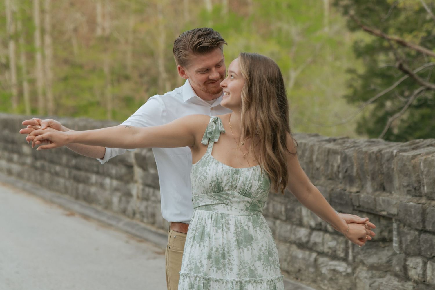 Smiling couple dancing playfully along a stone wall path lined with budding spring trees.