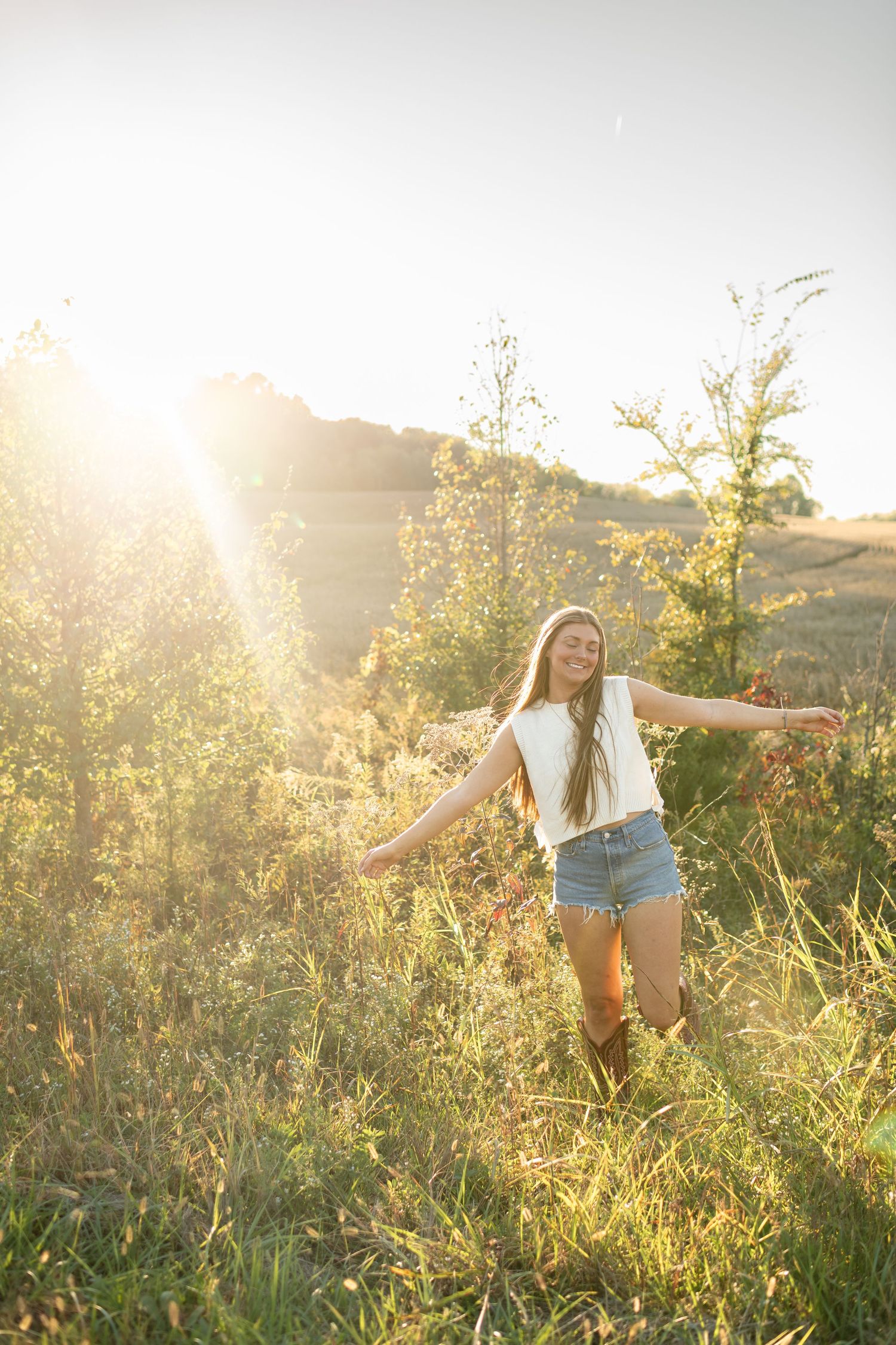 Young woman with long hair in white top and denim shorts stands in golden sunlit meadow with arms outstretched.