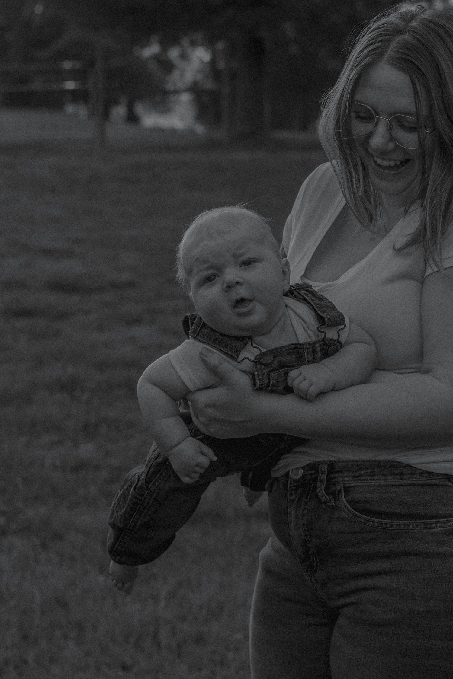Black and white photo of a smiling mother holding her baby boy outdoors in a park.