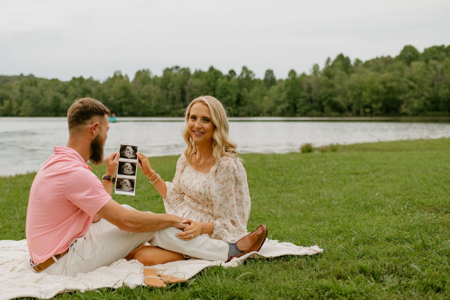 Couple sits on blanket by lake sharing ultrasound photos during a maternity announcement session.