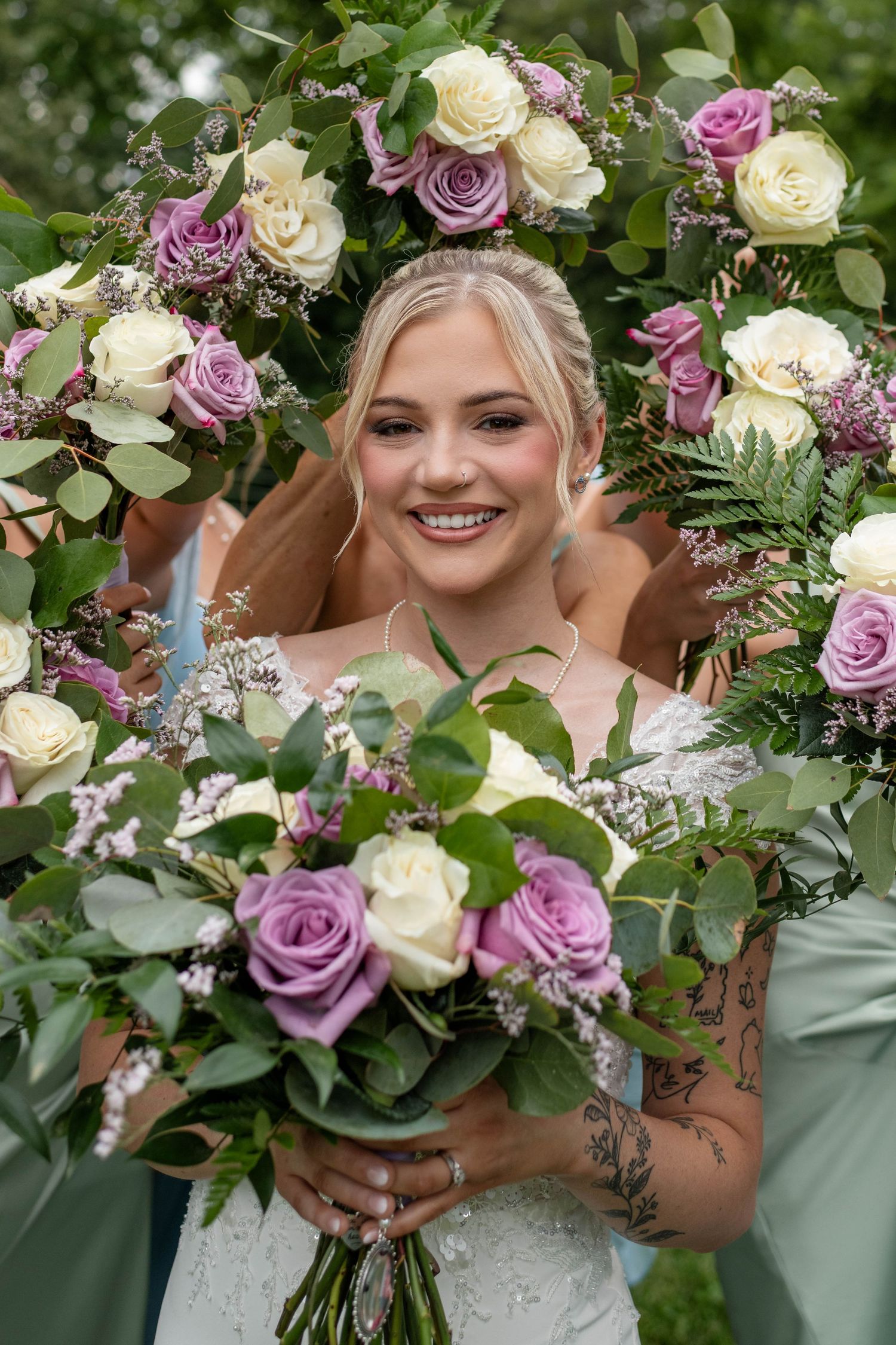 Smiling bride framed by purple and white rose bouquets held by bridesmaids in sage green dresses.