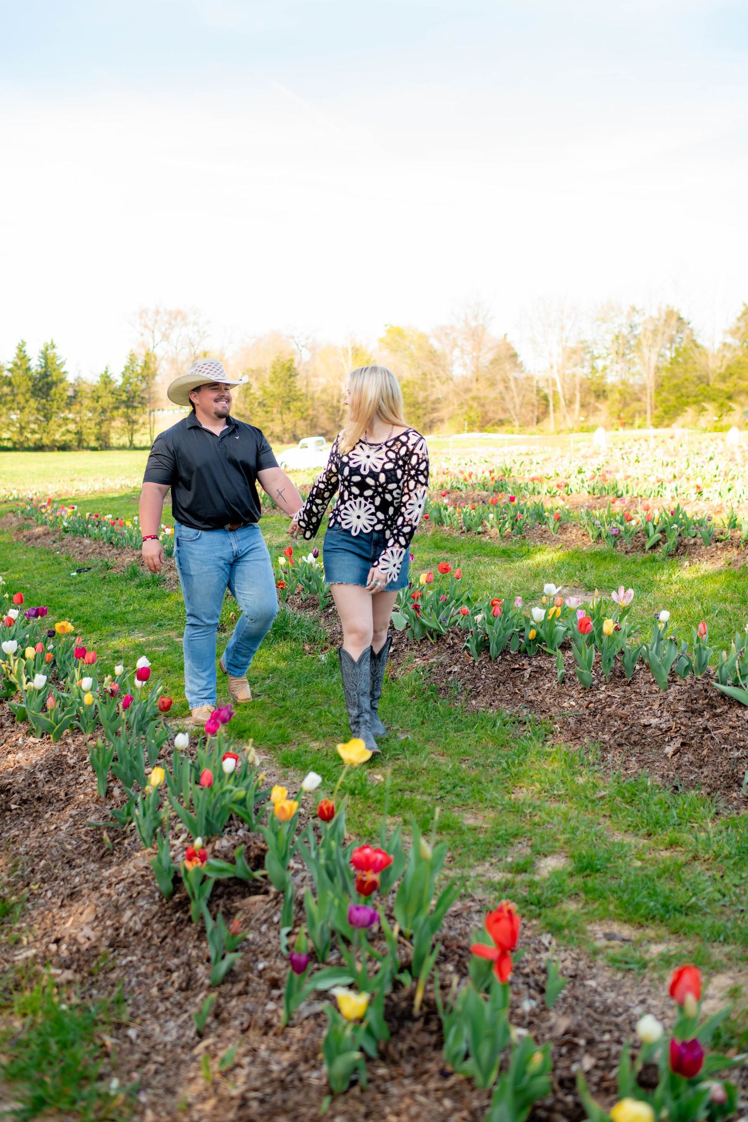 Couple walking through colorful tulip fields at golden hour, man in cowboy hat and woman in floral top.
