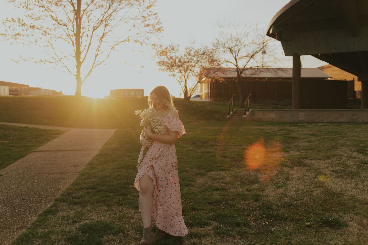 Woman in floral maxi dress holding flowers walks toward golden sunset with lens flare in open field.