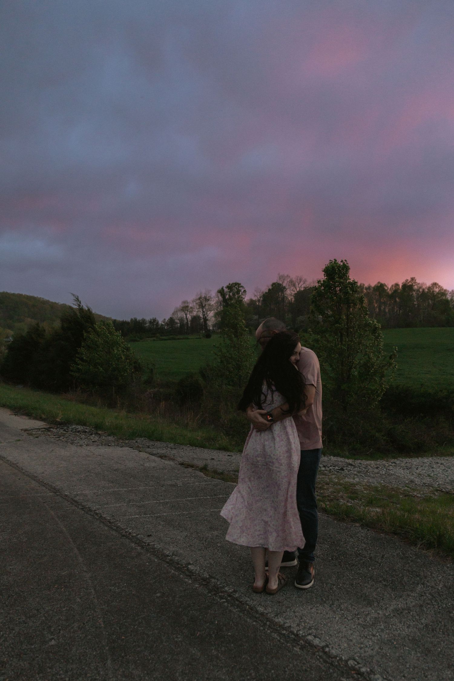 Romantic couple embracing on a rural road at sunset with vibrant pink and purple sky over green fields.