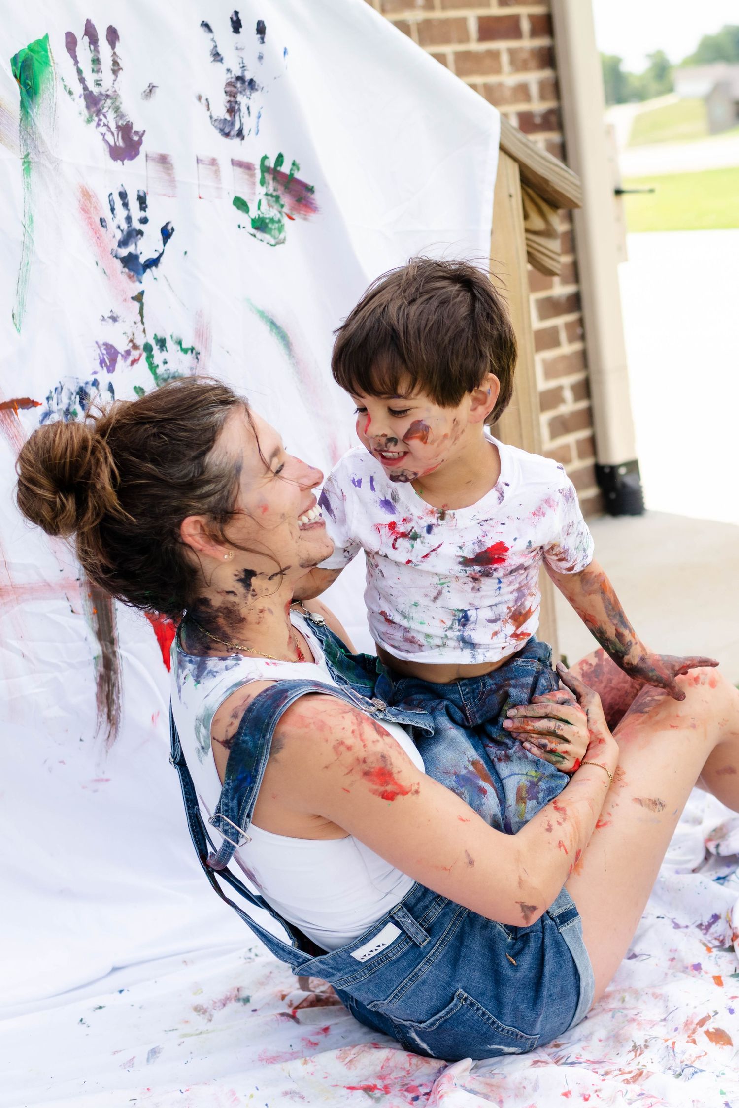 Laughing mother and paint-covered toddler sitting together in front of colorful handprint art.