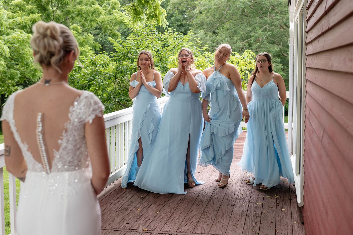 Bridesmaids in light blue gowns react with excitement during first look with bride on wooden deck.
