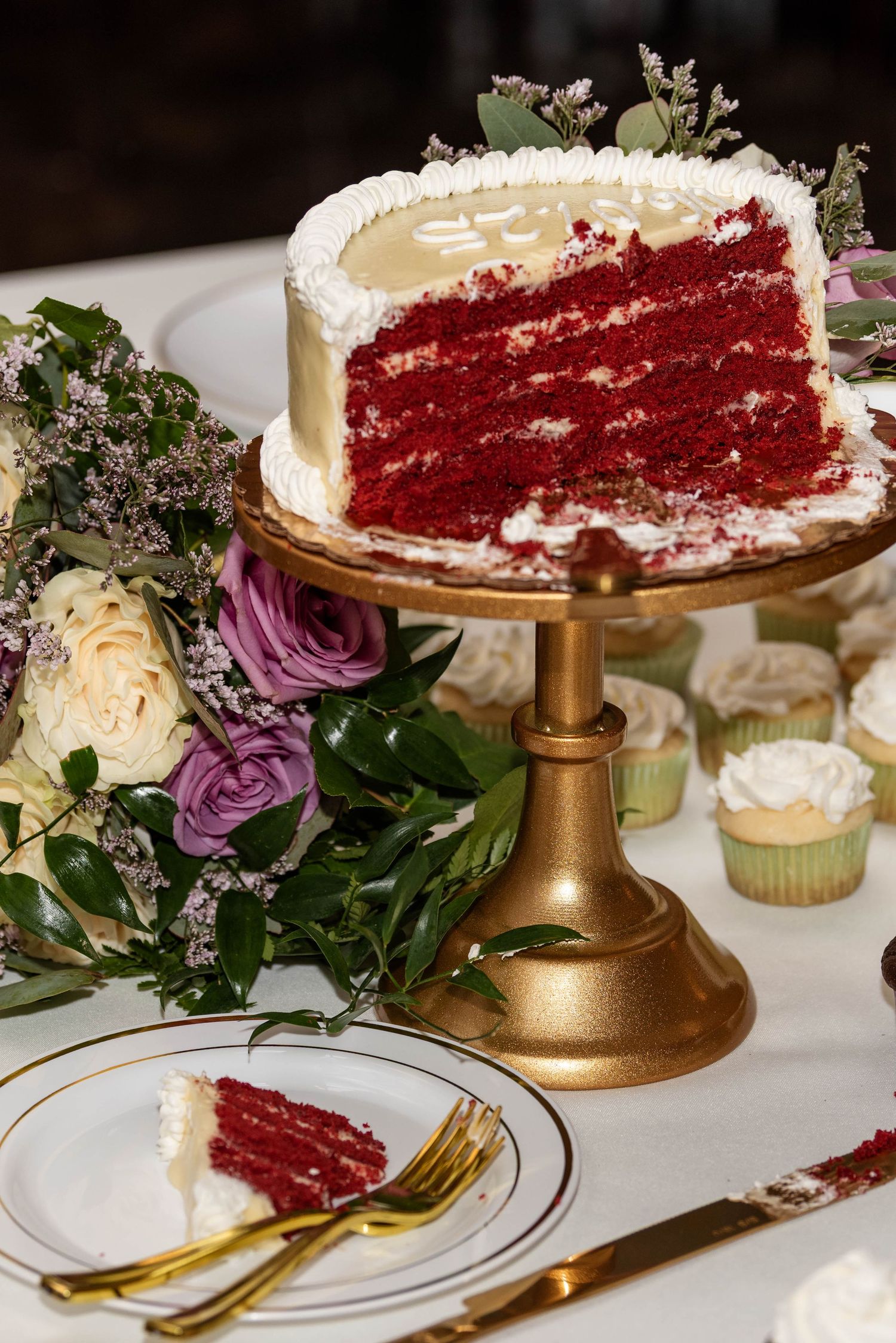 Sliced red velvet wedding cake on a gold pedestal stand surrounded by flowers and cupcakes.