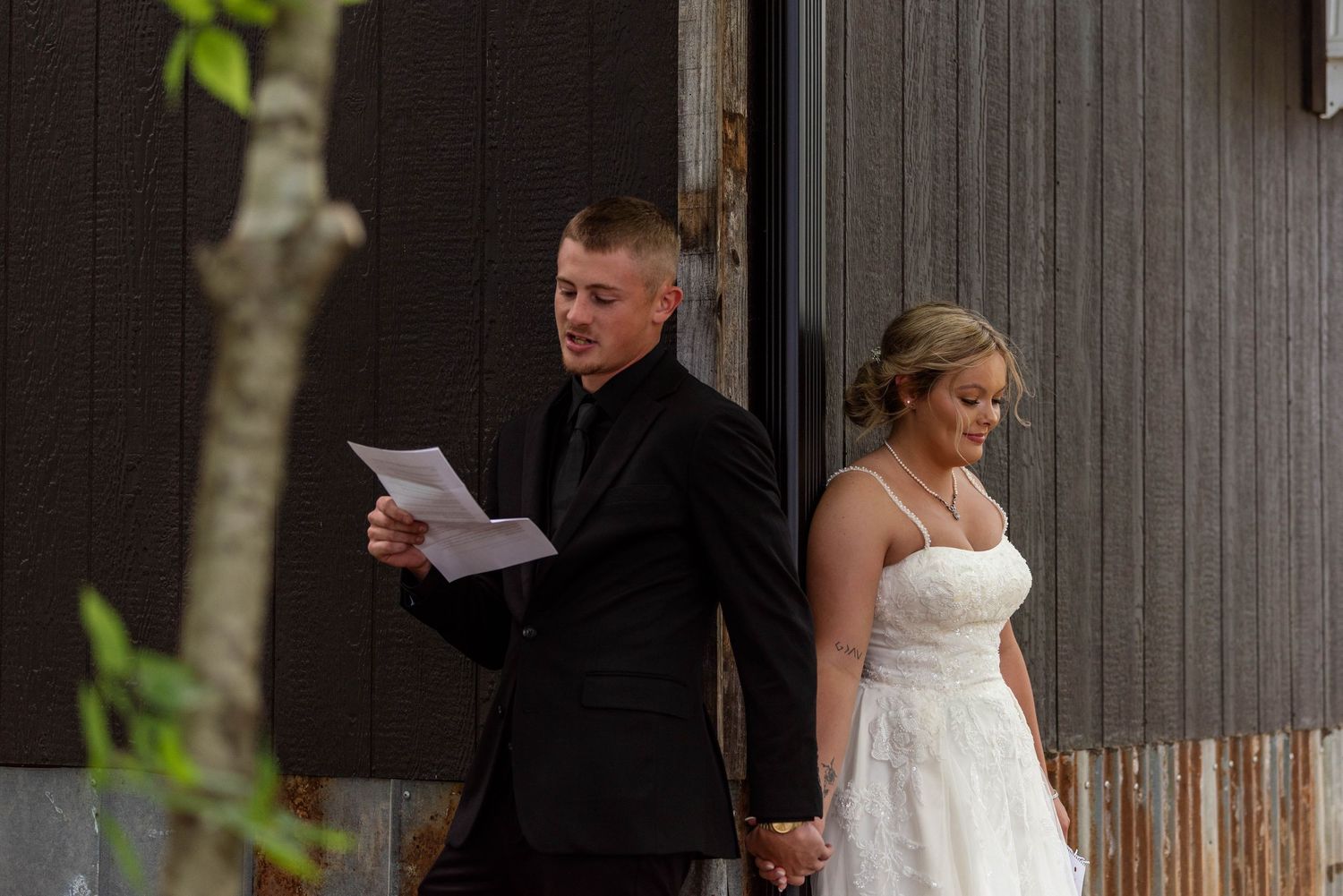 Groom reads vows from paper while holding hands with bride outside a rustic dark barn wall.