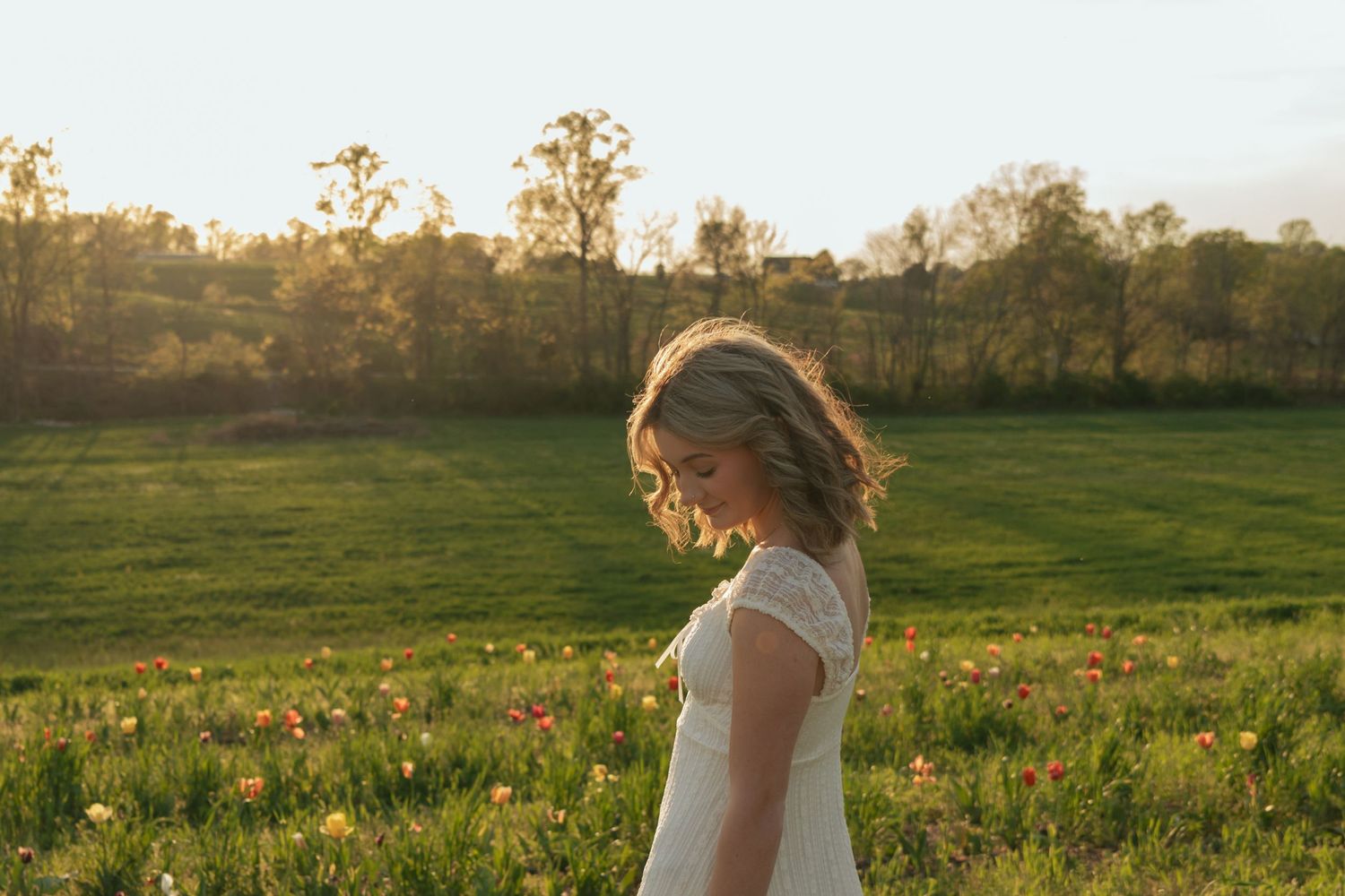 Woman in white lace dress standing in golden sunlit meadow with wildflowers at dusk.