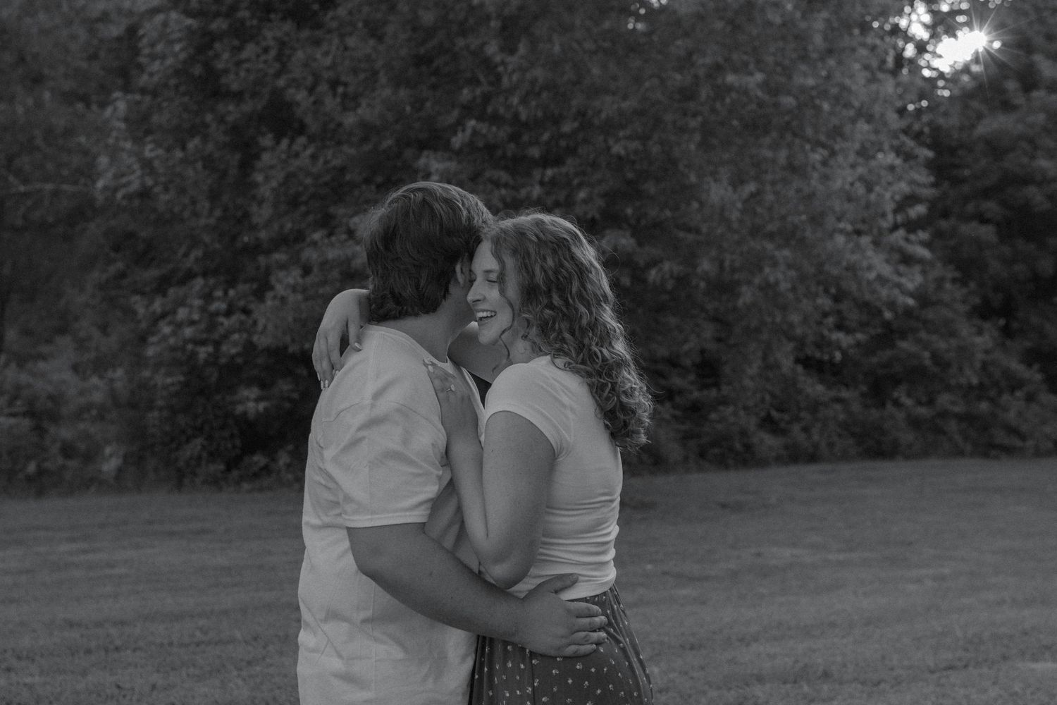 Black and white photo of happy engaged couple embracing and laughing together in an outdoor park setting.