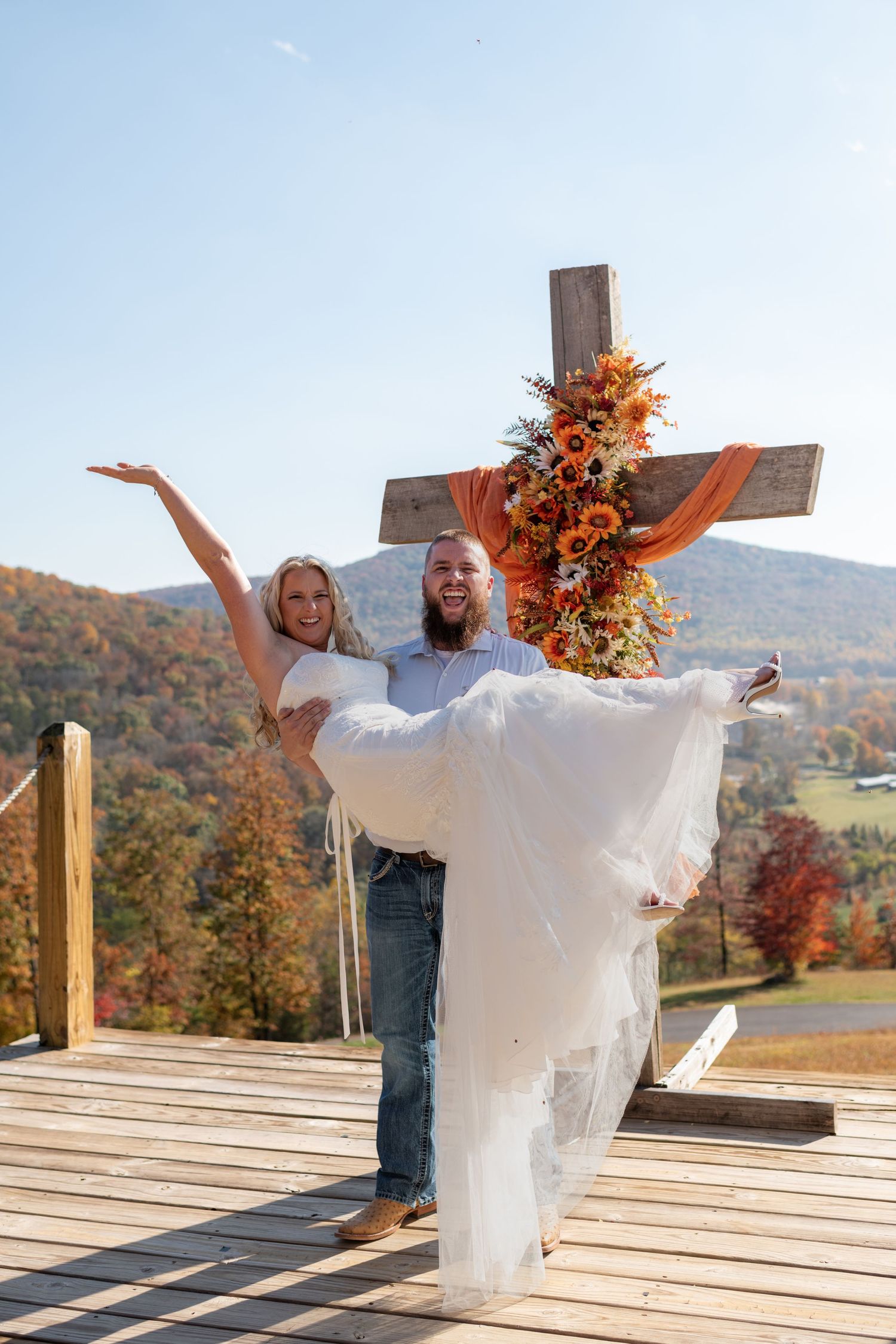 Joyful groom carries bride in white dress before a floral cross with mountain autumn backdrop.