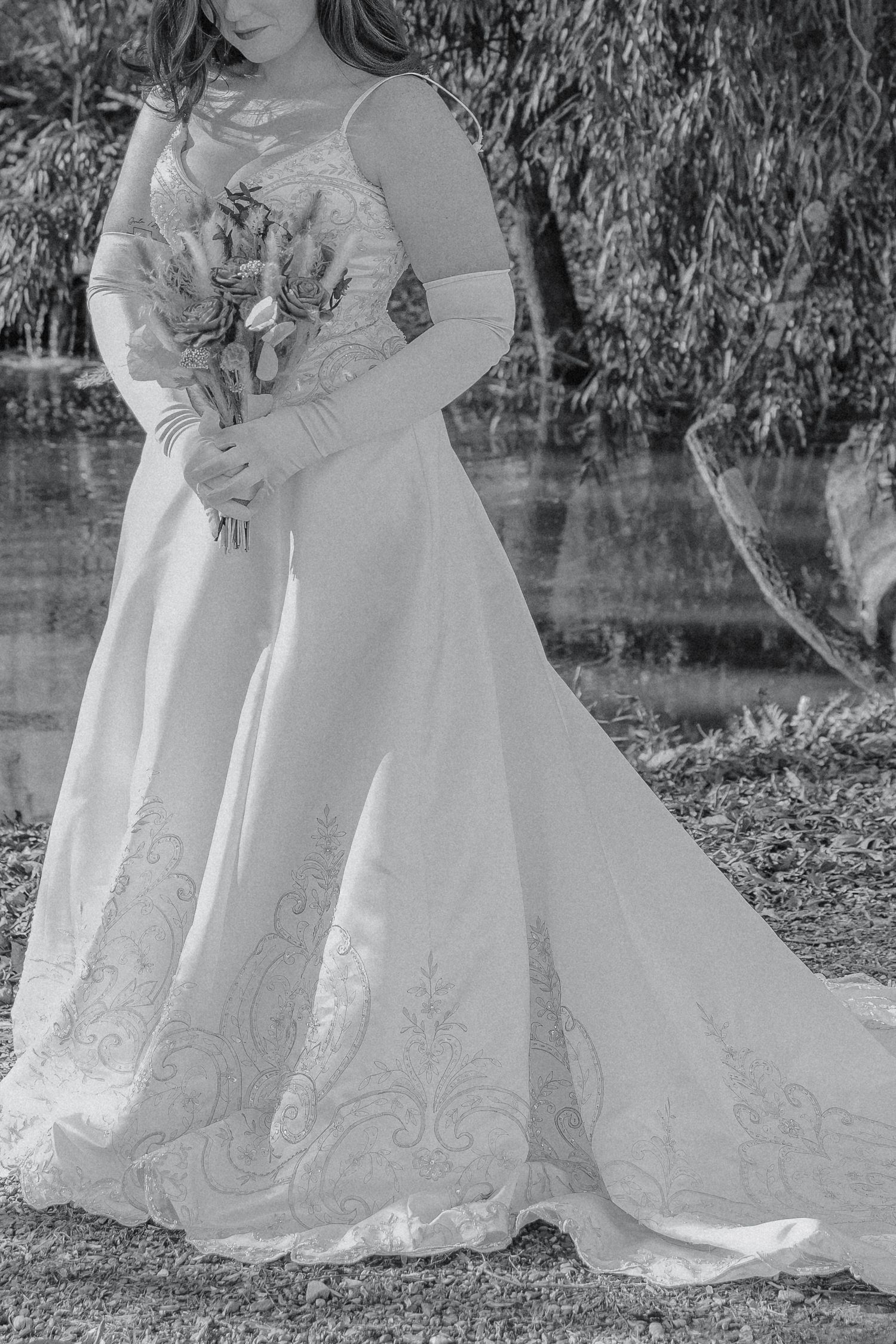 Black and white photo of bride in lace gown holding a bouquet near a willow tree.