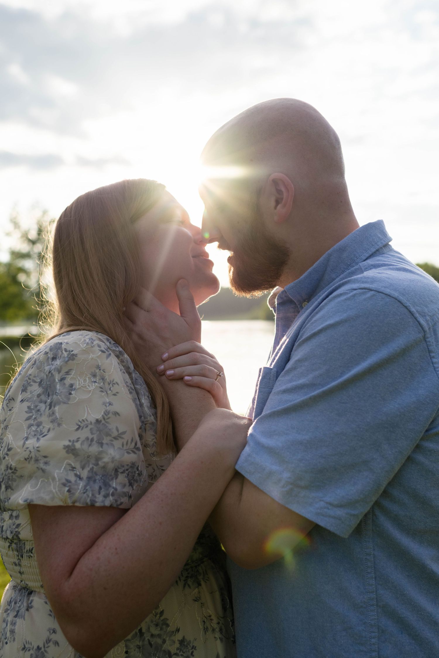 Romantic couple sharing a sunset kiss with golden sun flare glowing between them near a lake.