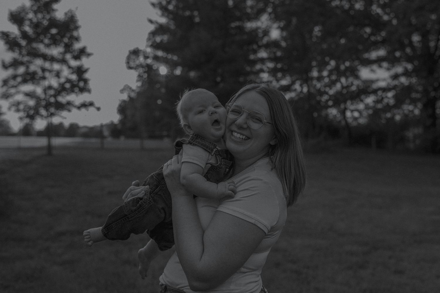 Black and white photo of a smiling mother holding her baby in an open field.