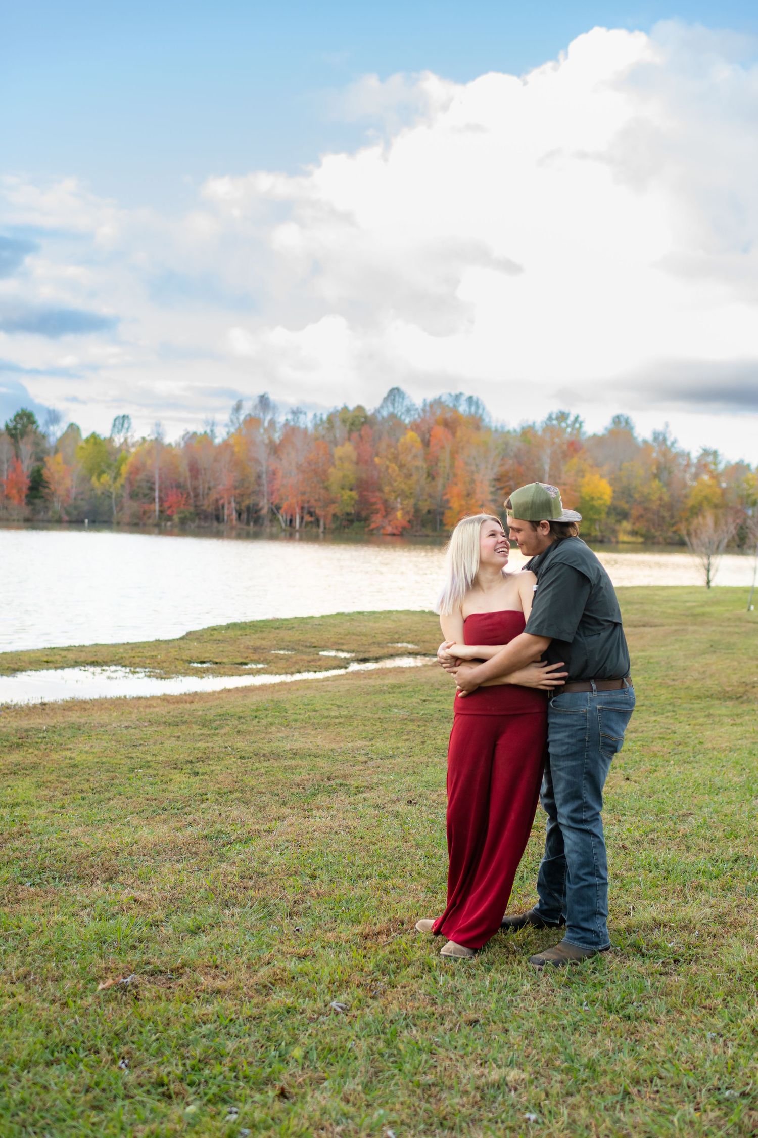 Couple standing close together by a lake surrounded by colorful fall foliage under a dramatic sky.