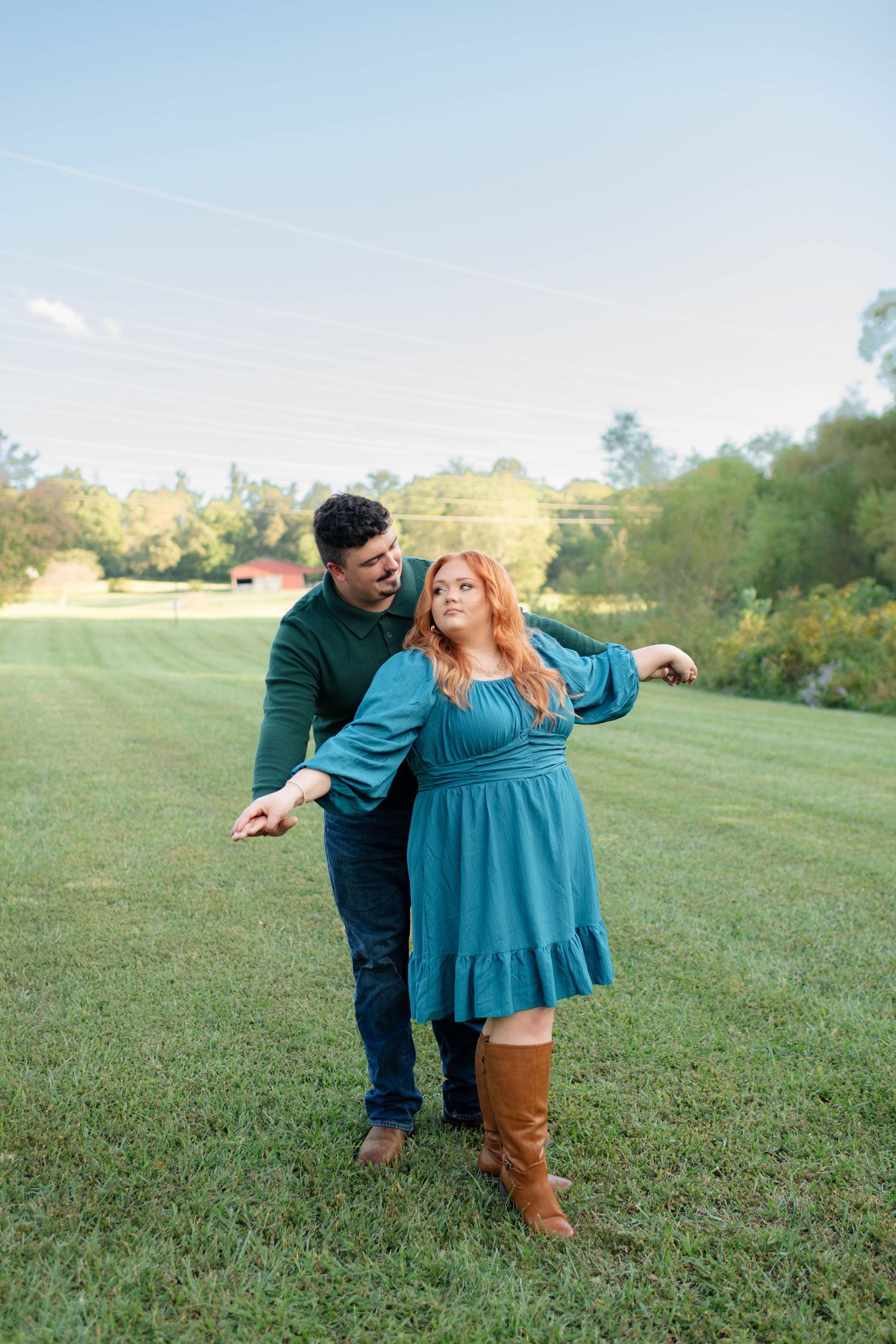 Couple dancing playfully on a green lawn with trees and a red barn visible in the background.