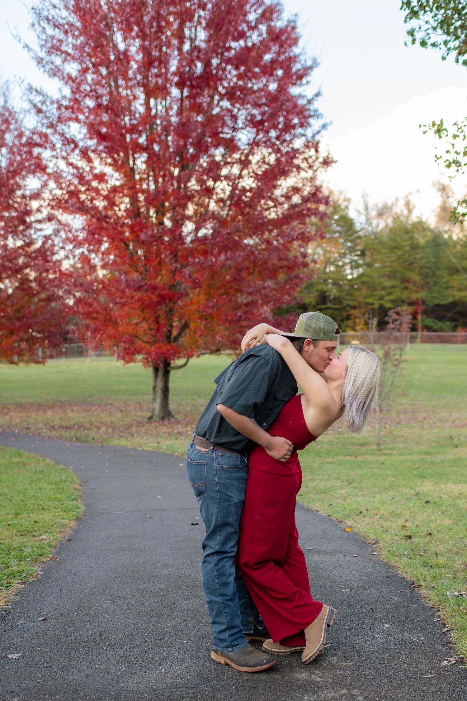 Couple sharing passionate kiss on pathway with vibrant red autumn tree in background.
