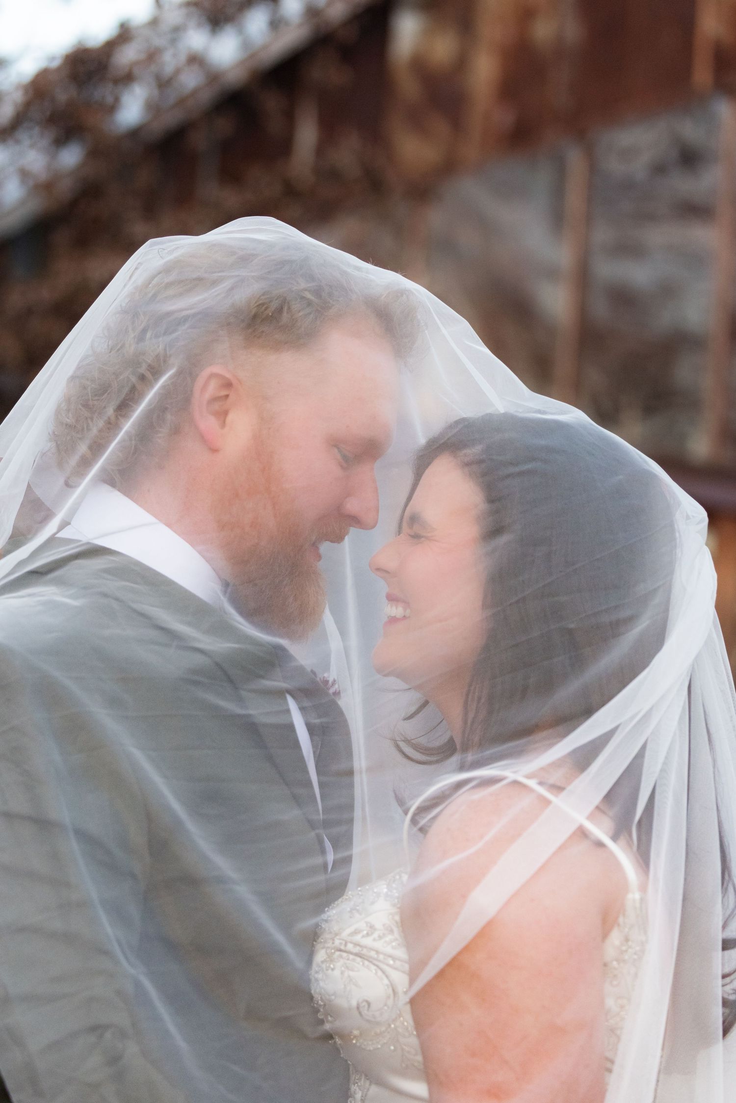Bride and groom share an intimate moment under a wedding veil, smiling at each other outdoors.