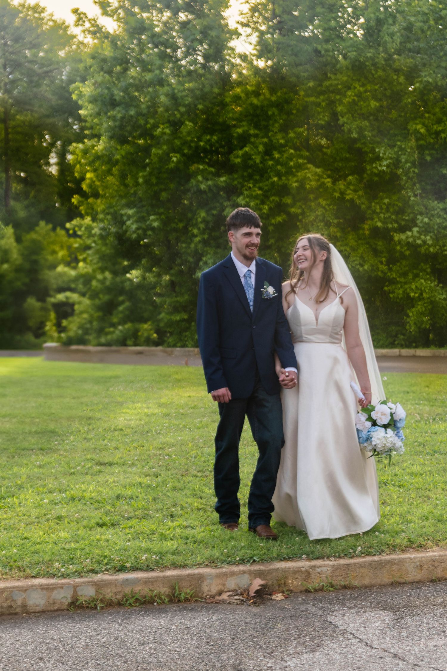 Bride and groom walk hand in hand on a sunlit path surrounded by green trees.
