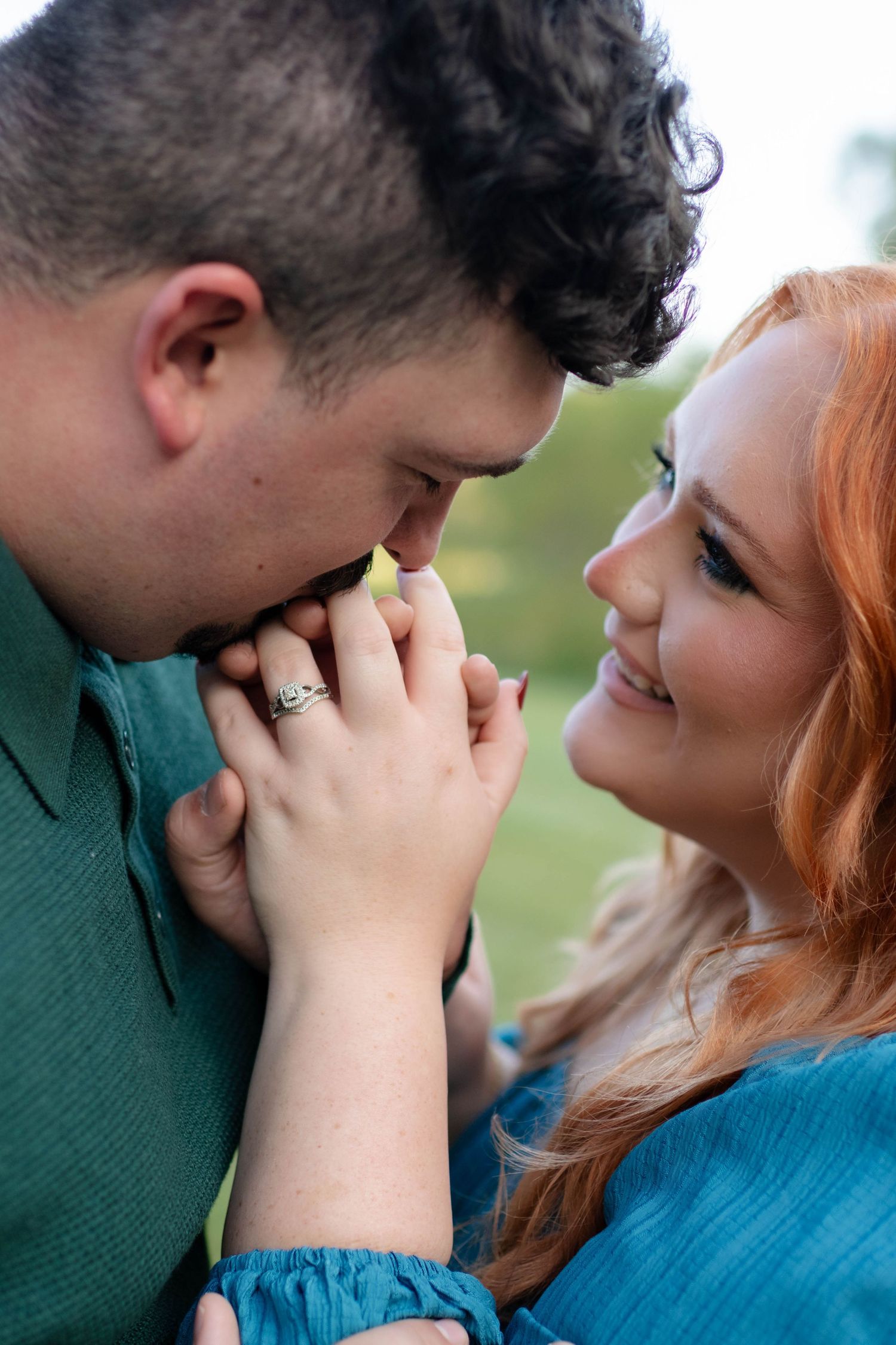 Man kissing woman's hand as she smiles warmly, showcasing an engagement ring in a tender outdoor moment.