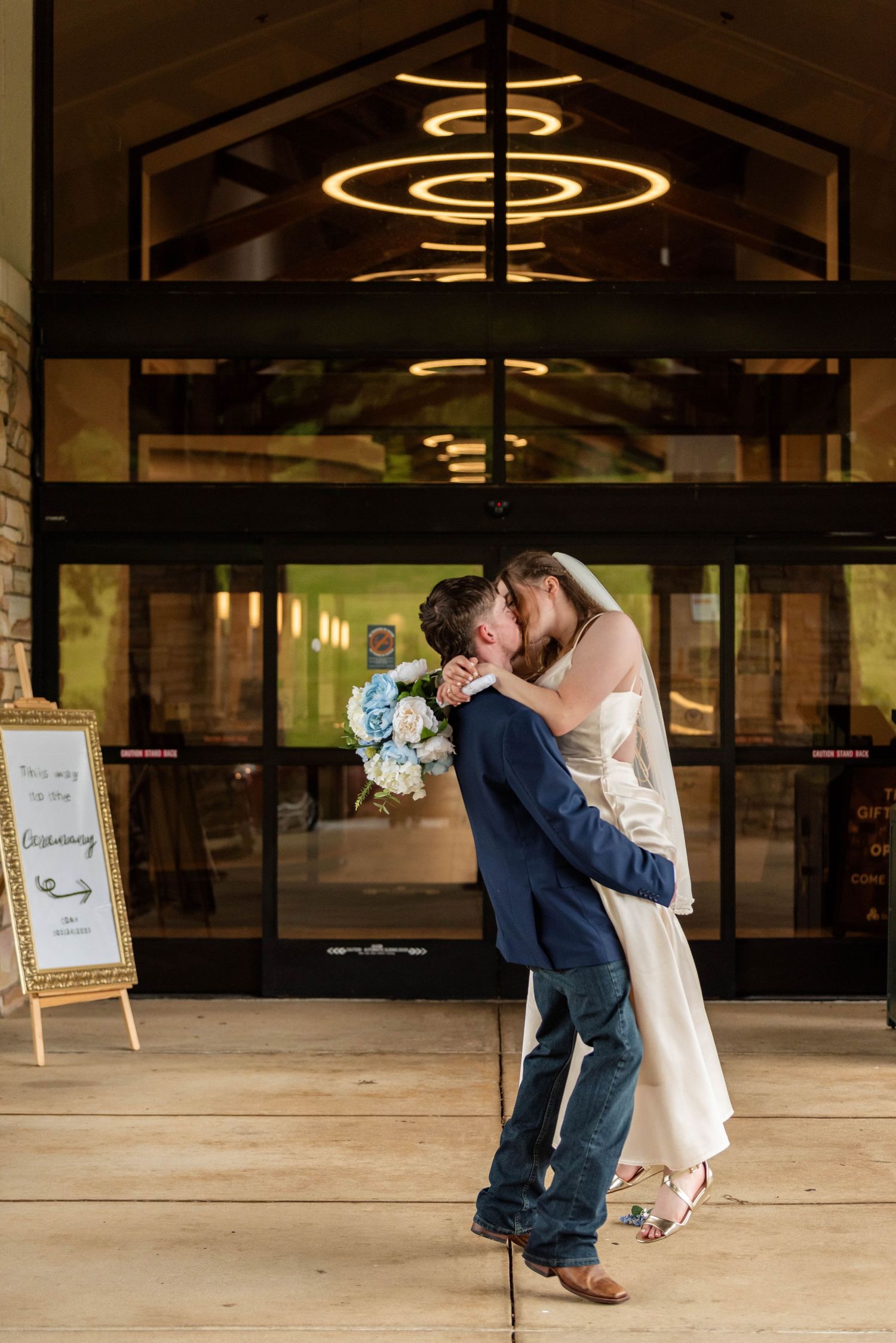 Groom lifts bride for a kiss outside venue entrance holding blue bouquet.