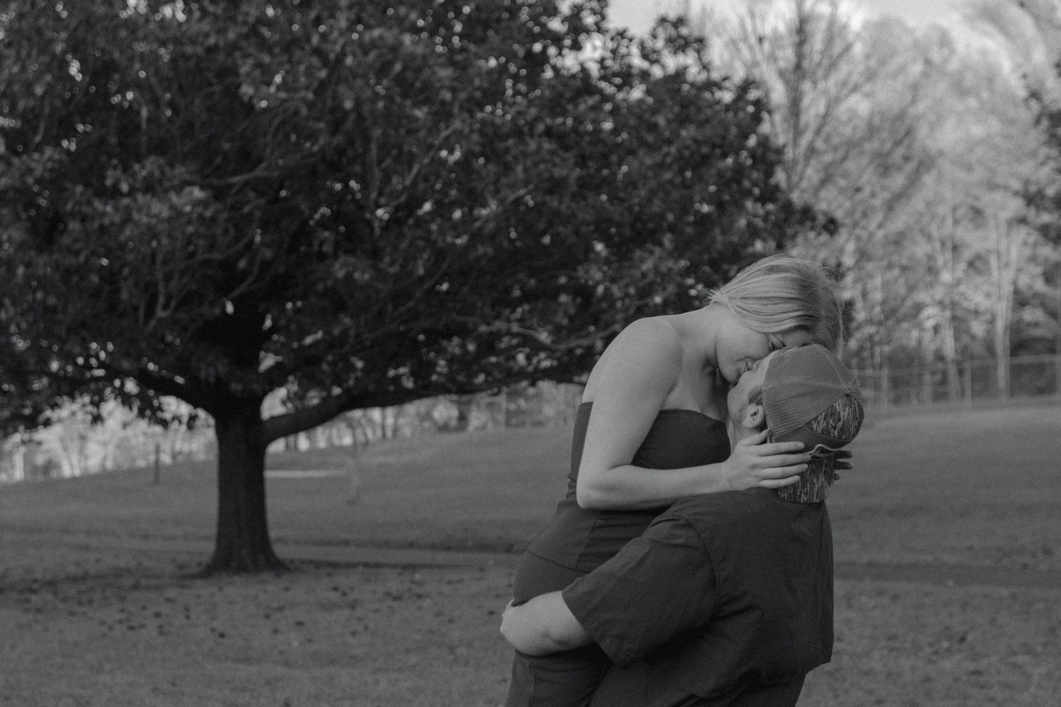 Black and white photo of couple embracing tenderly in open park with large tree backdrop.