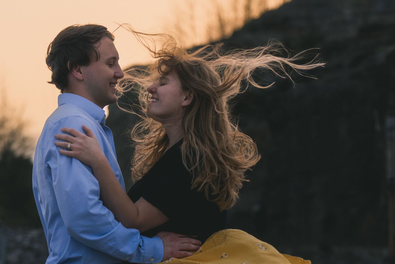 Joyful couple embracing at sunset with woman's hair blowing in the wind against golden sky.
