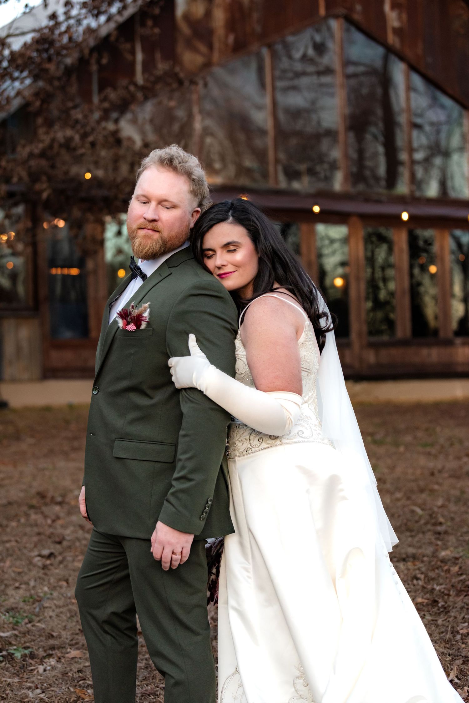 Bride in white gown hugging groom in green suit outside a rustic wedding venue.