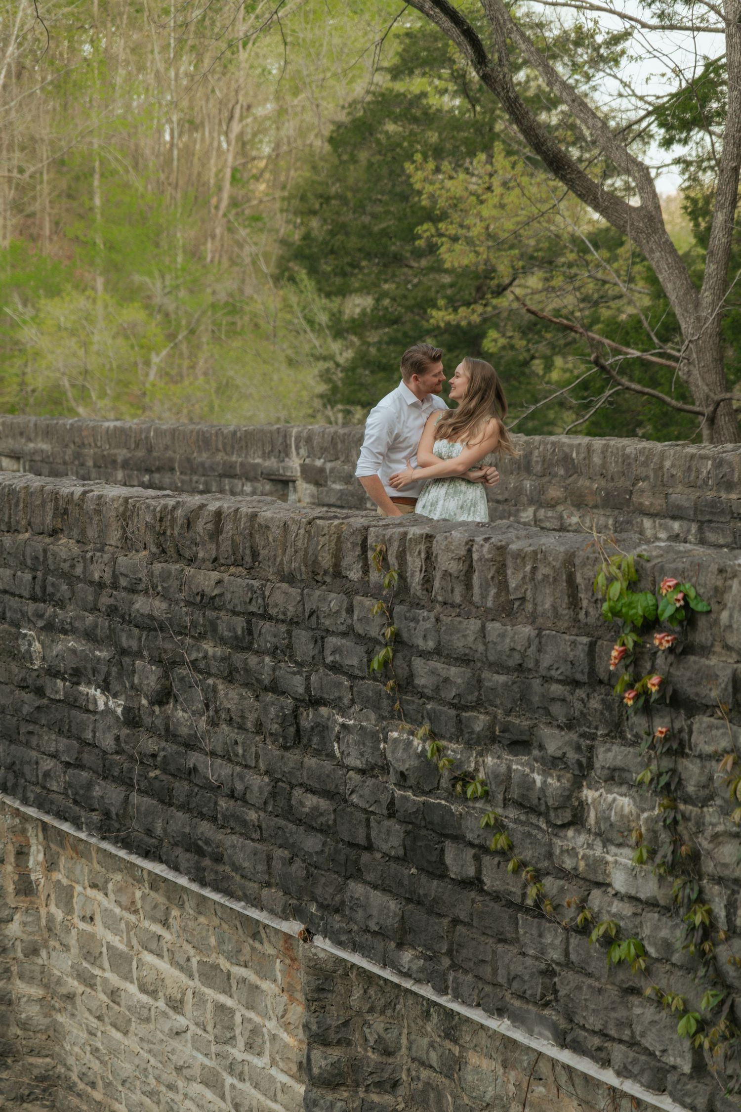 Romantic couple embracing and kissing atop an old stone bridge wall surrounded by lush green trees.