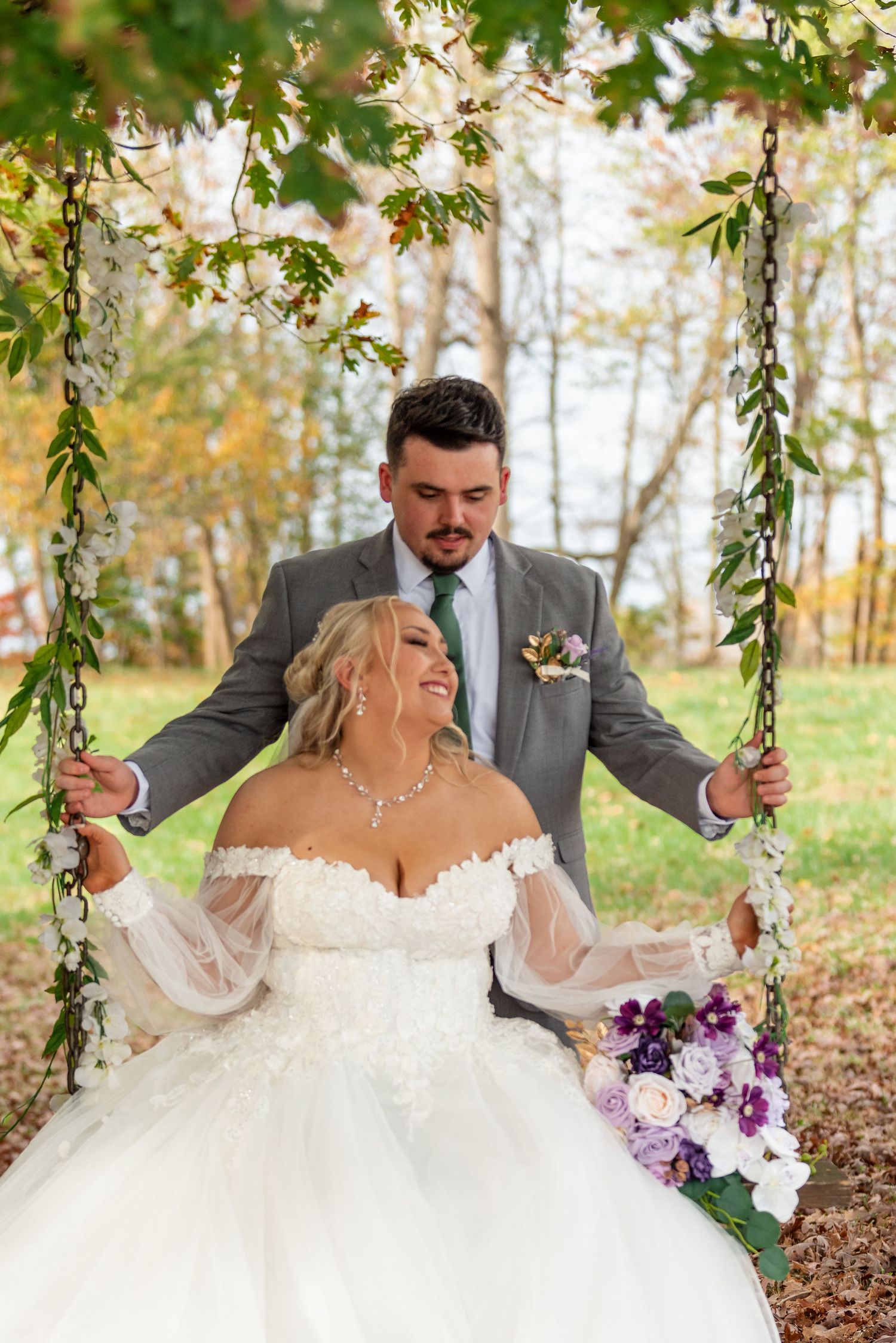 Smiling bride sits on a floral swing holding purple bouquet as groom stands behind her outdoors.