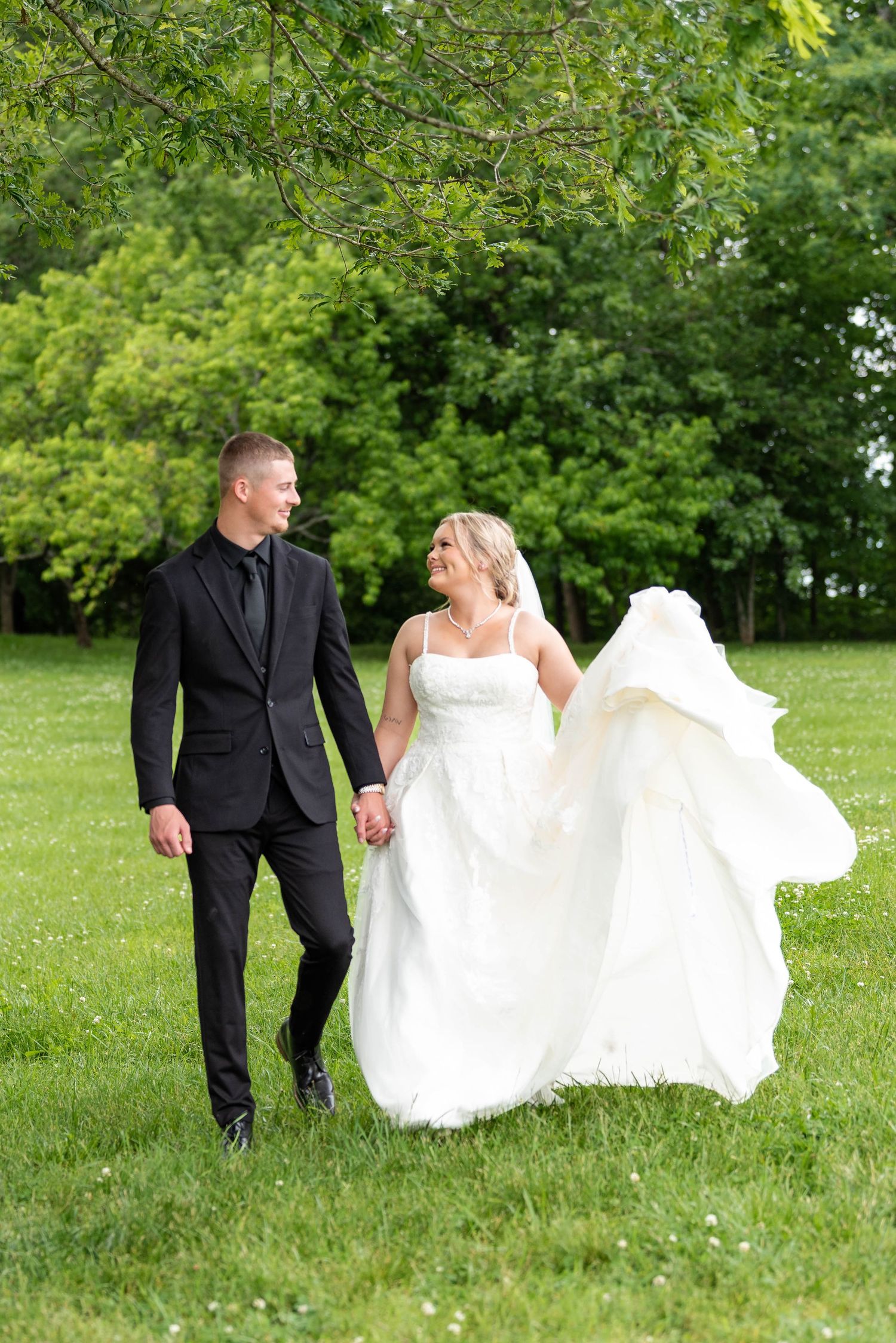 Bride and groom walking hand in hand through green meadow, white dress flowing in the breeze.
