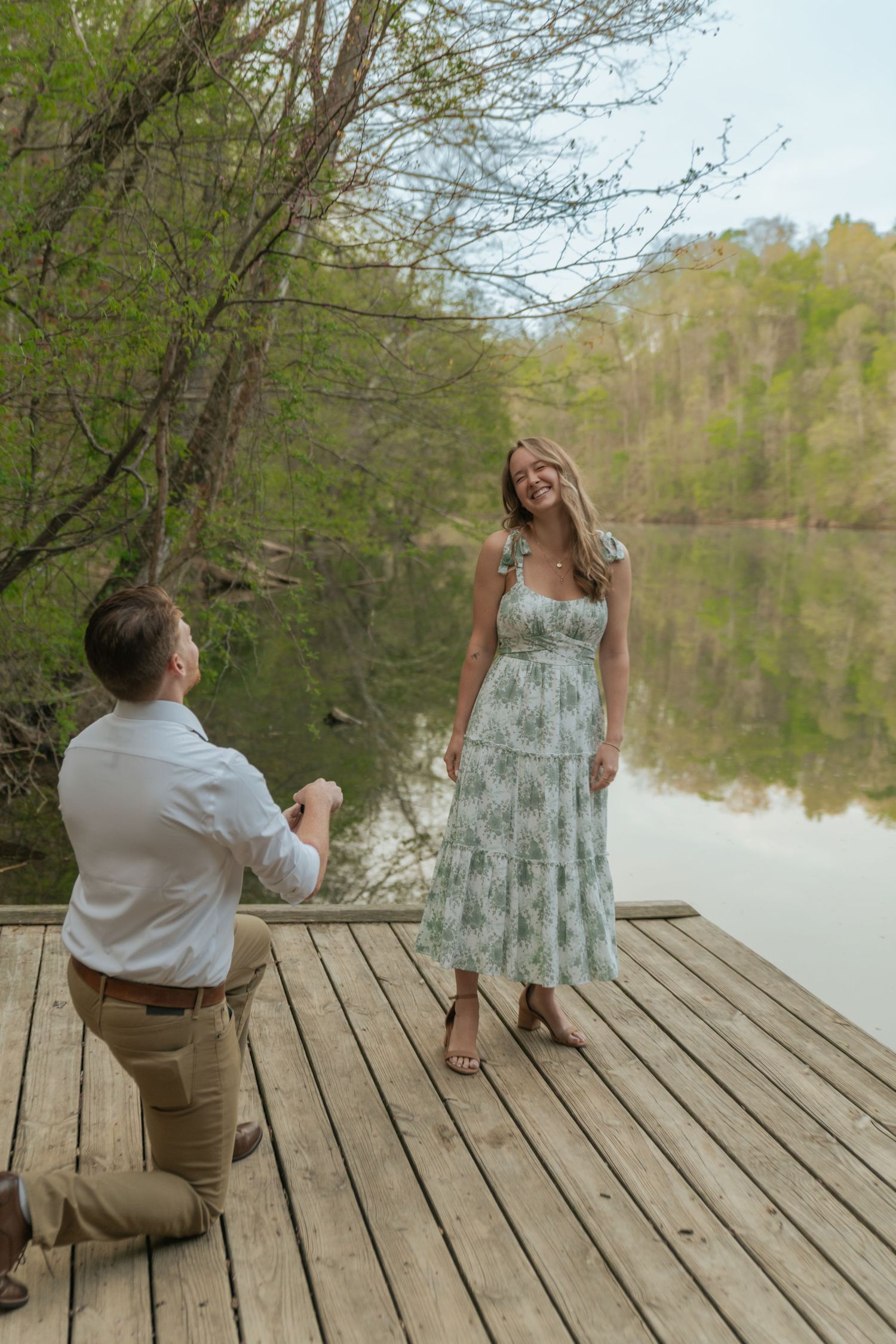 Man proposing on one knee to a woman on a wooden dock by a serene lake surrounded by lush green trees.